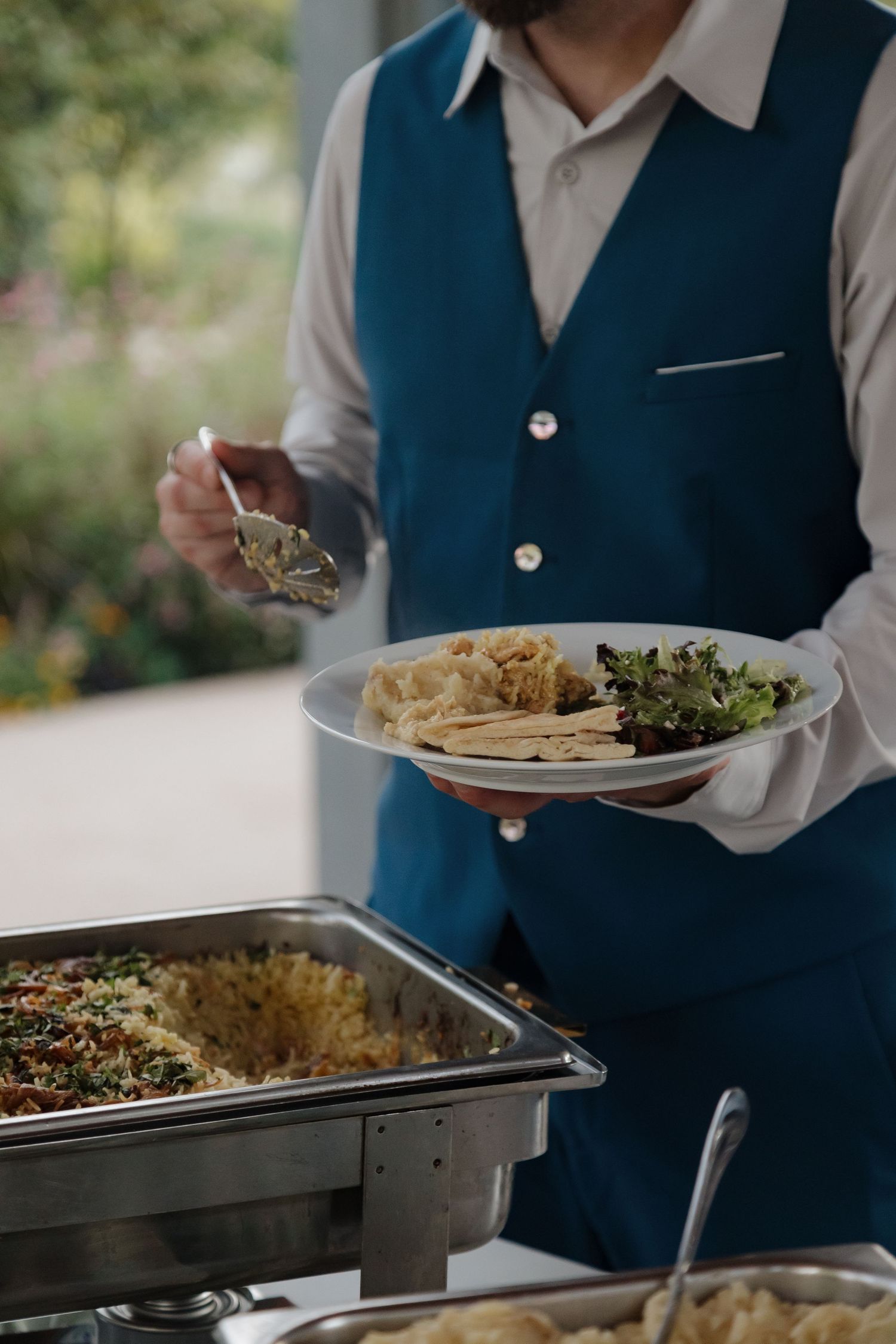 Catering staff member in blue vest serves food from buffet chafing dishes at wedding reception.