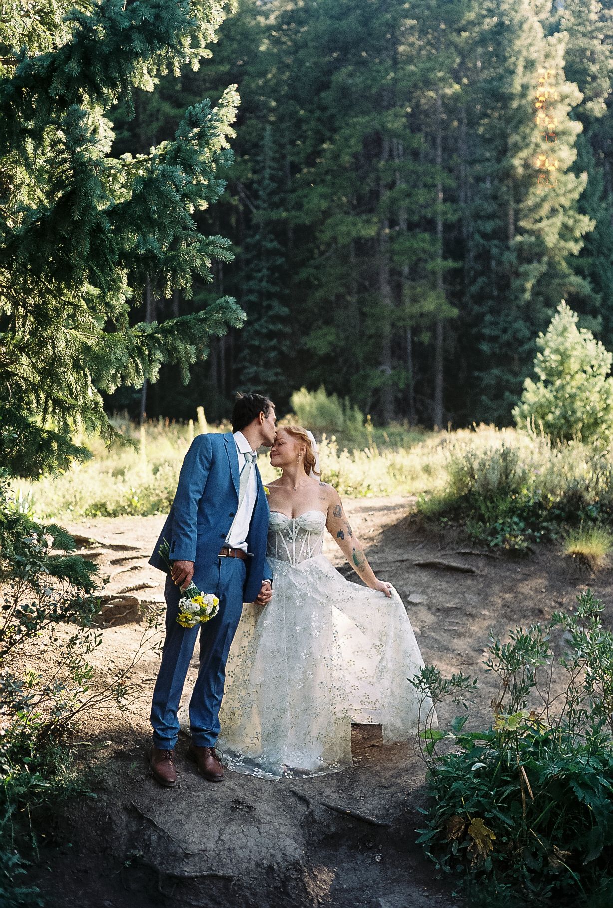 Bride and groom share a tender forehead touch on a sunlit forest path among tall pine trees.