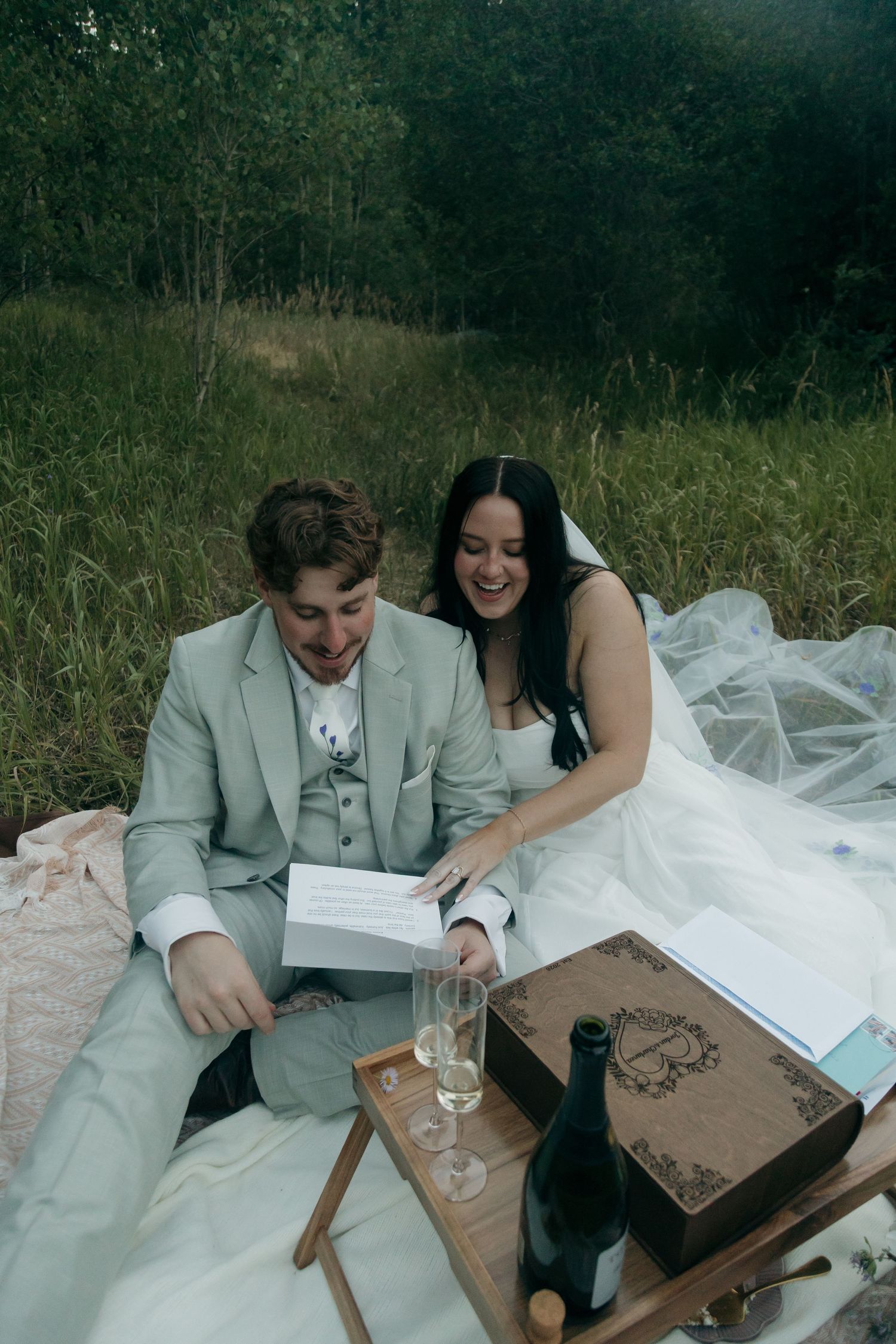 Laughing bride and groom reading vow books on picnic blanket in forest meadow with champagne glasses.