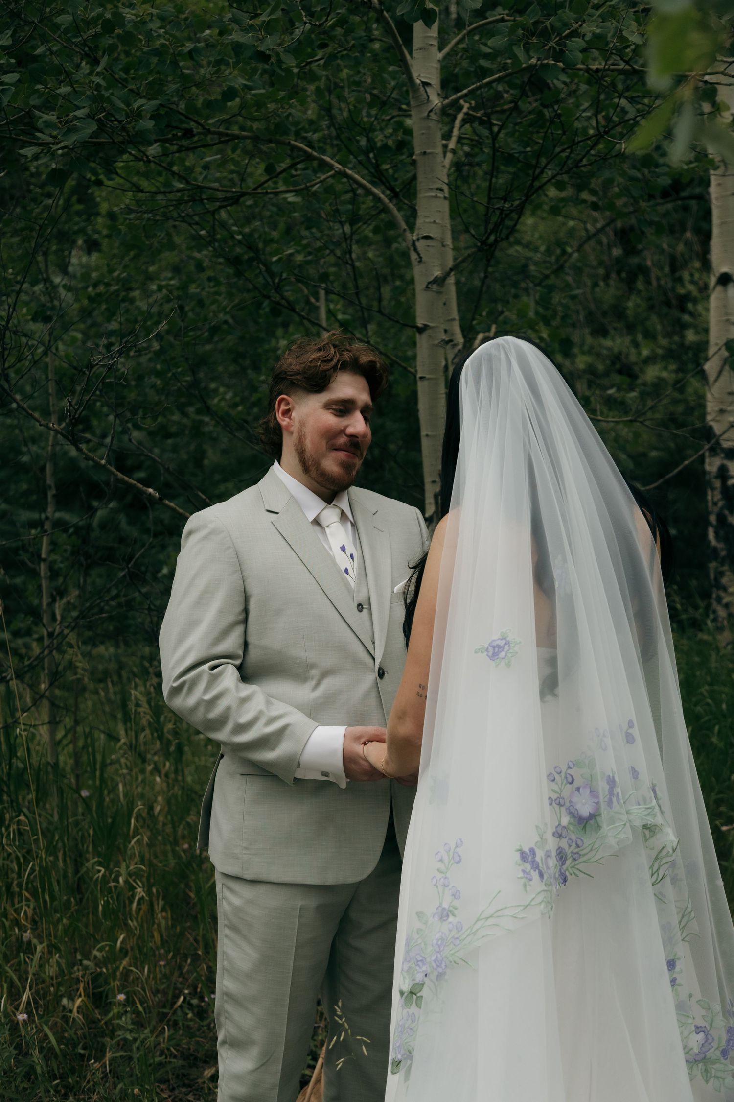 Groom in light gray suit smiles at bride wearing embroidered floral veil during outdoor woodland ceremony.