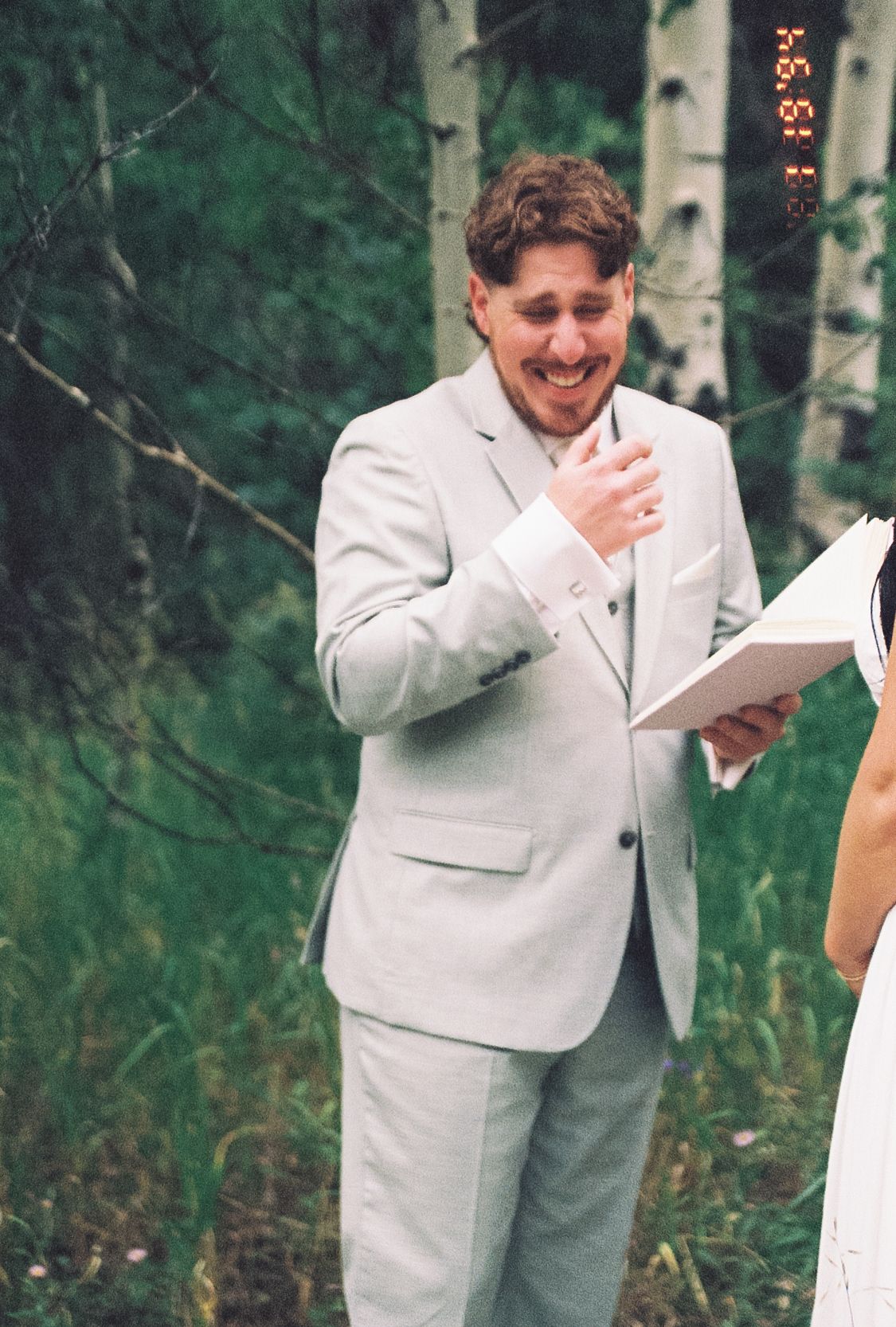 Emotional groom in grey suit laughs joyfully during outdoor forest wedding ceremony.