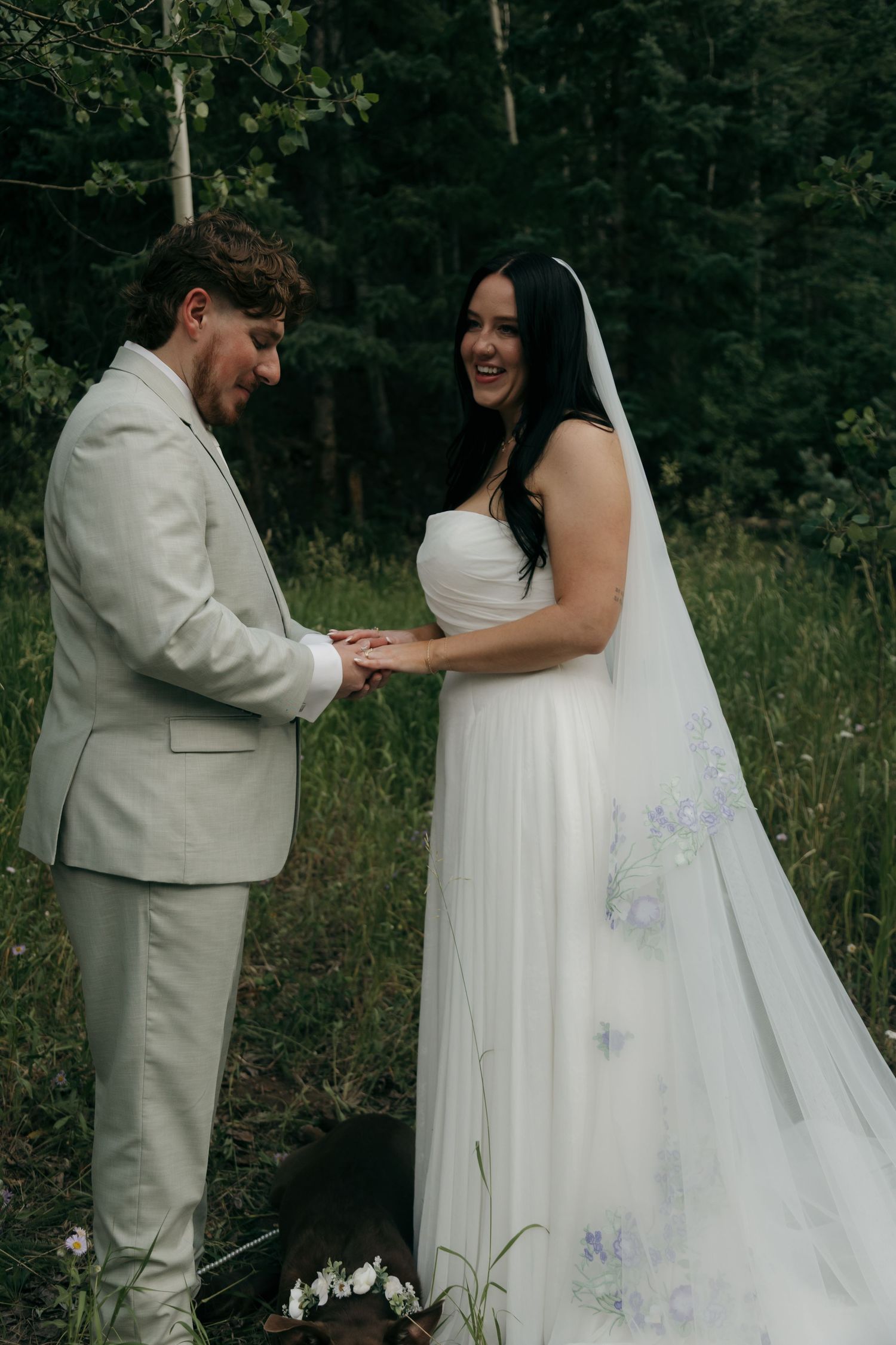 Smiling bride in white gown with floral veil holds hands with groom in beige suit in forest clearing.
