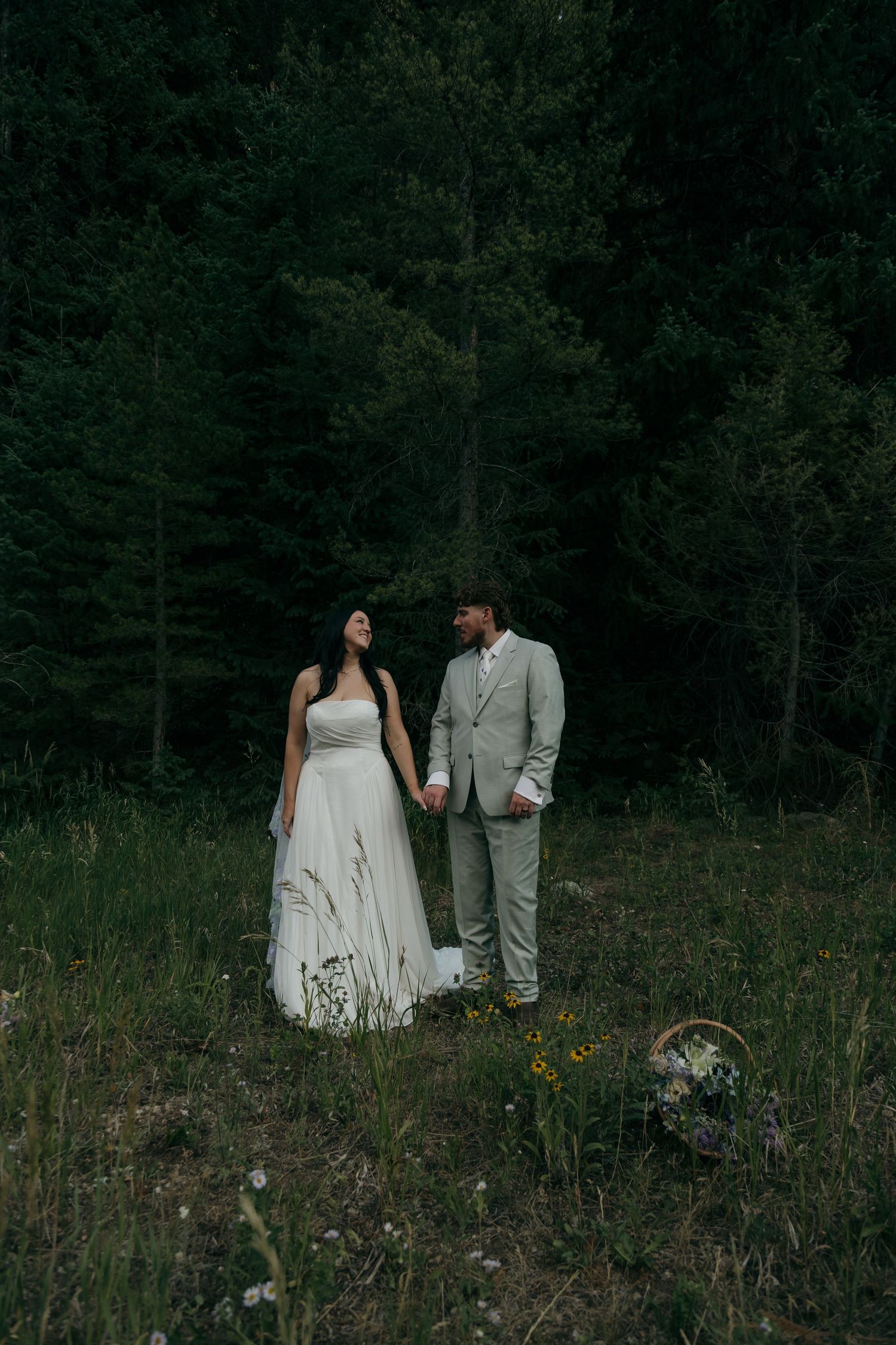 Bride and groom hold hands gazing at each other in dark forest clearing with wildflower bouquet nearby.
