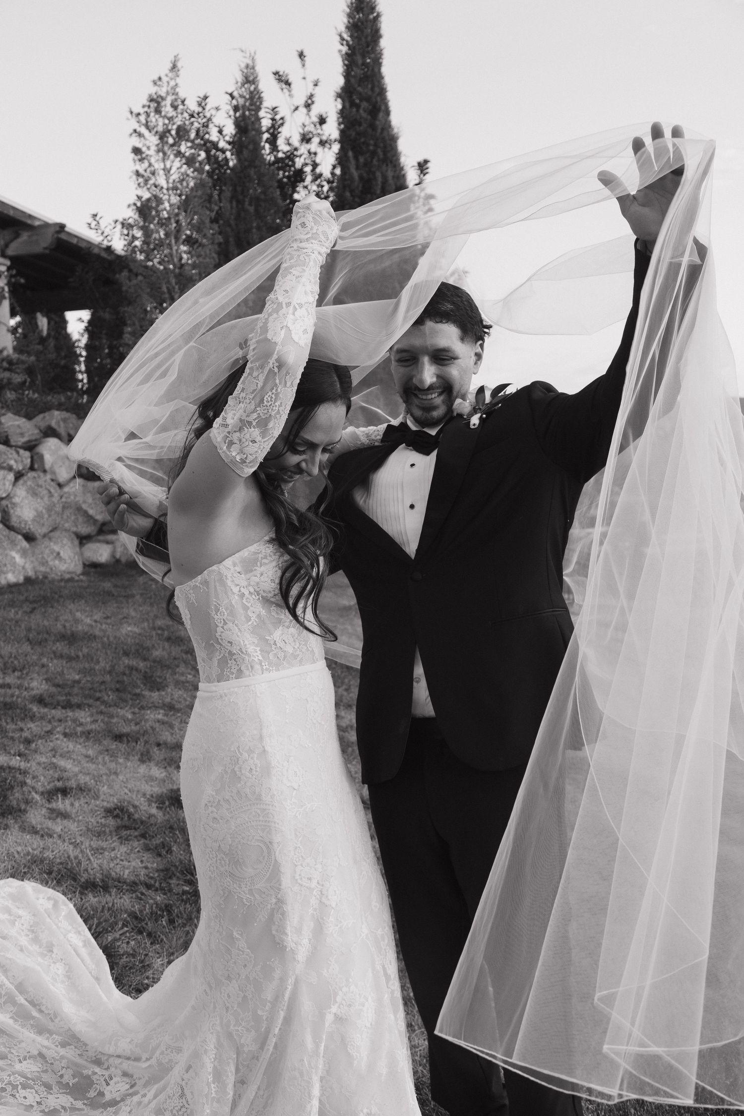 Black and white photo of a smiling groom lifting bride's veil outdoors at their wedding ceremony.