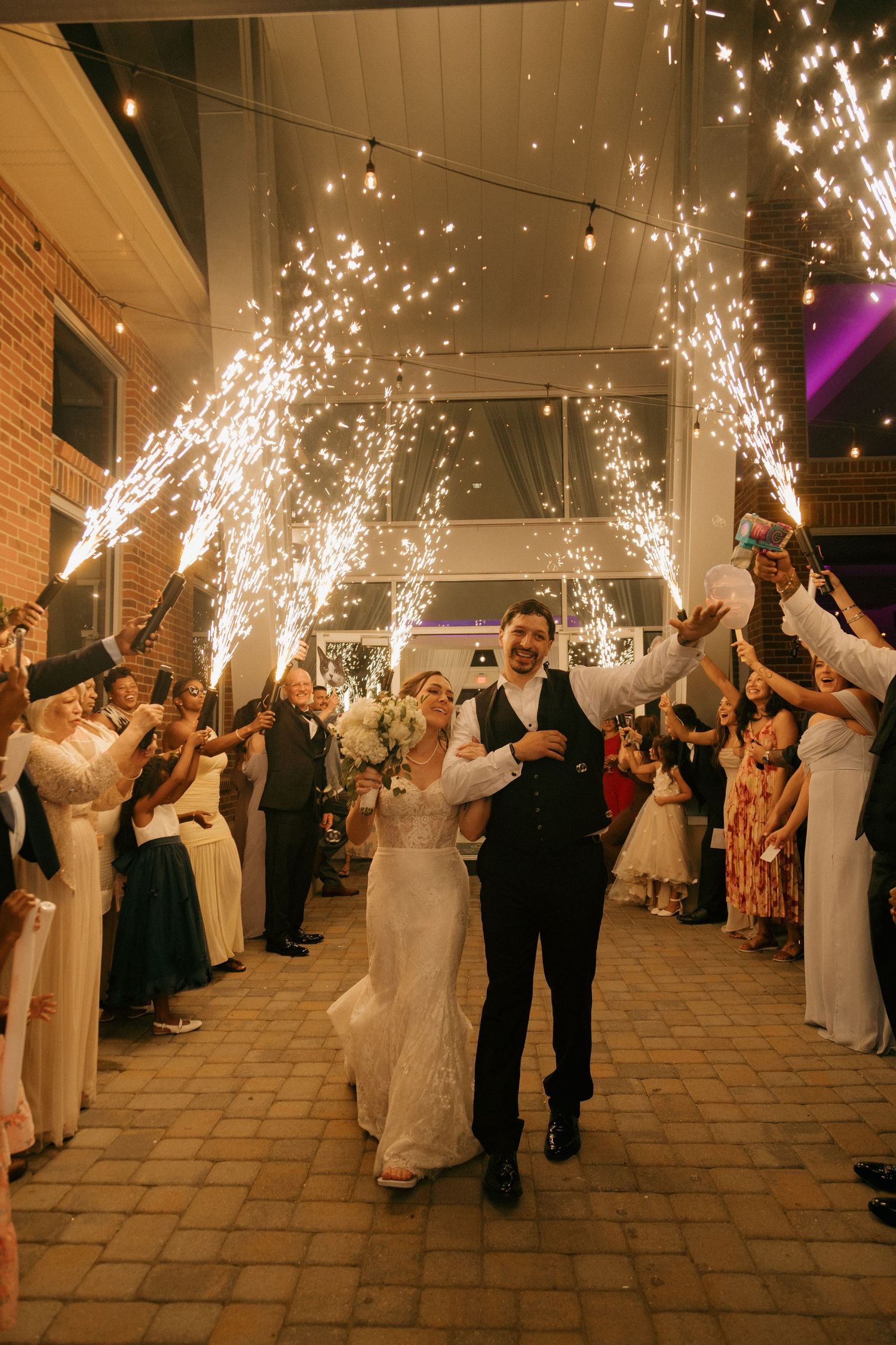 Smiling newlyweds walk through sparkling cold fireworks tunnel during wedding grand exit.