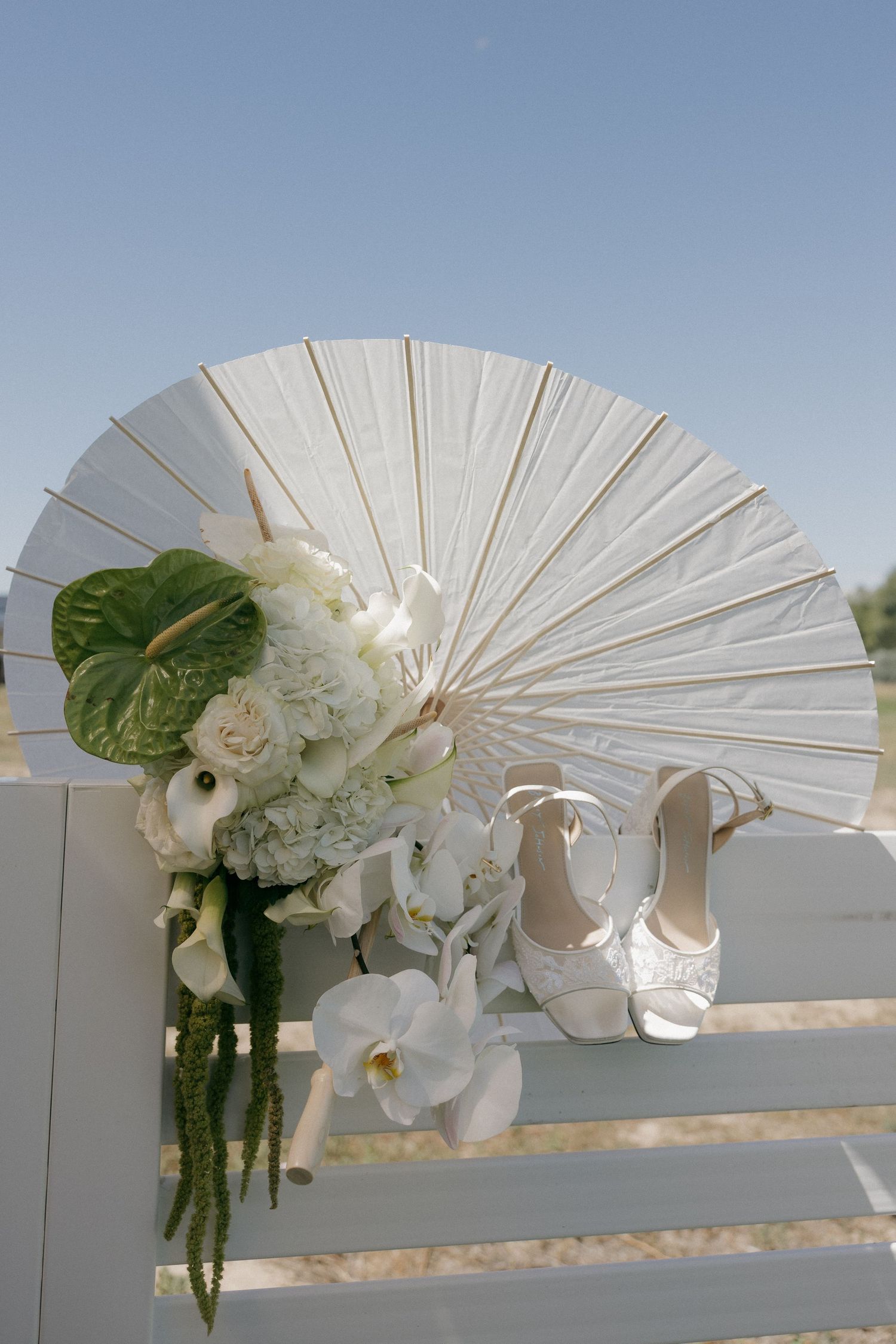 White wedding parasol, bridal bouquet with orchids, and strappy heels displayed on white fence under clear blue sky.