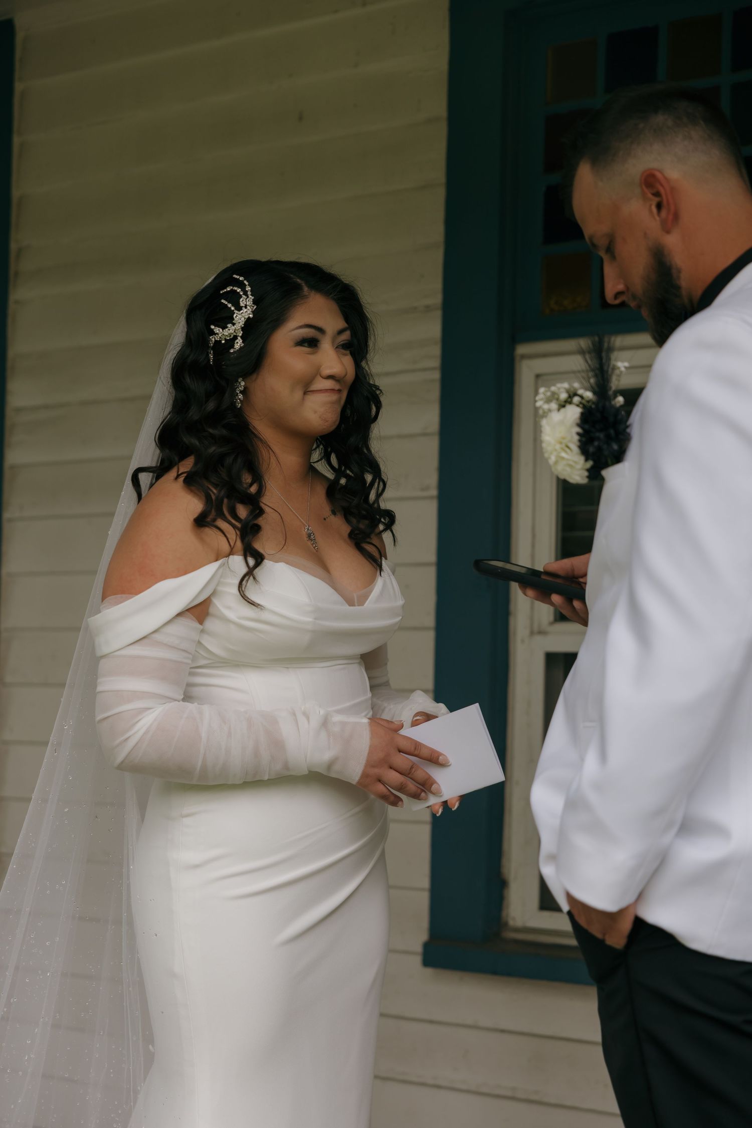 Bride in off-shoulder white gown smiles emotionally as groom reads wedding vows from phone on porch.