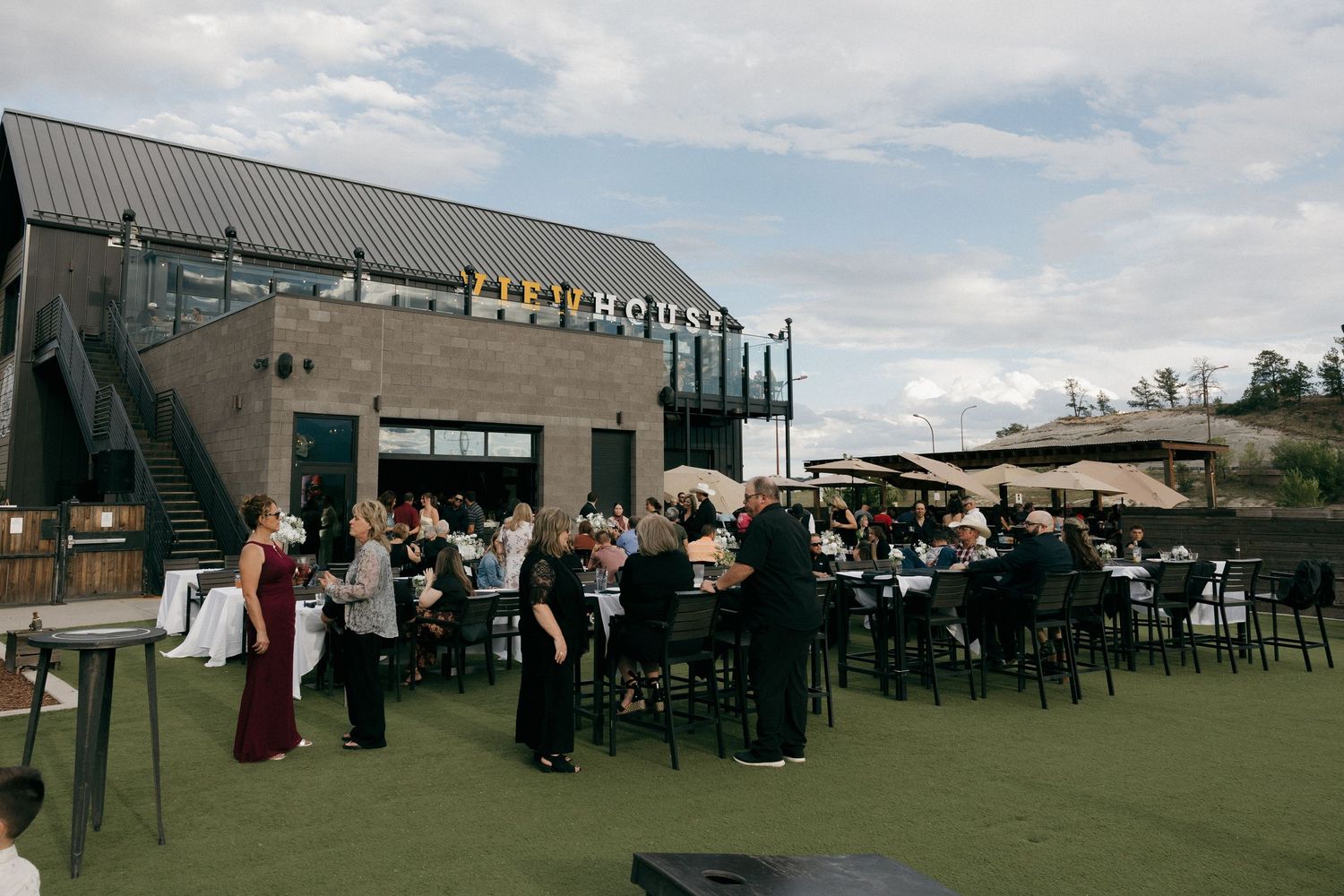 Outdoor rooftop event venue with guests dining at black tables on artificial turf under a partly cloudy sky.