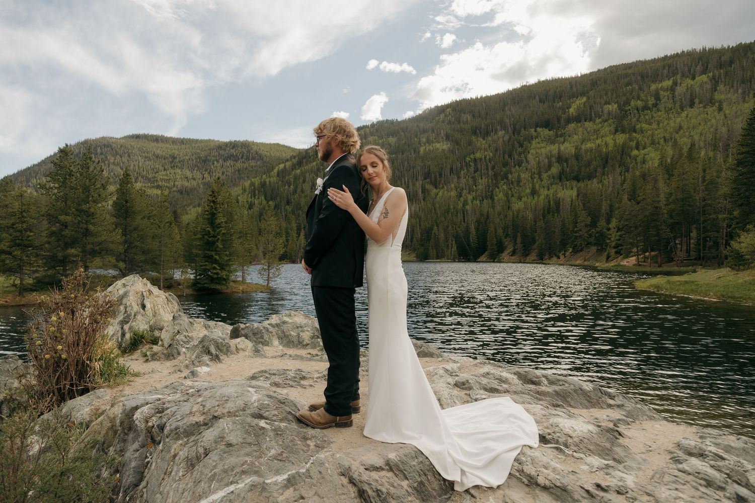 Bride and groom embracing on rocky lakeside with mountain forest backdrop during outdoor wedding ceremony.