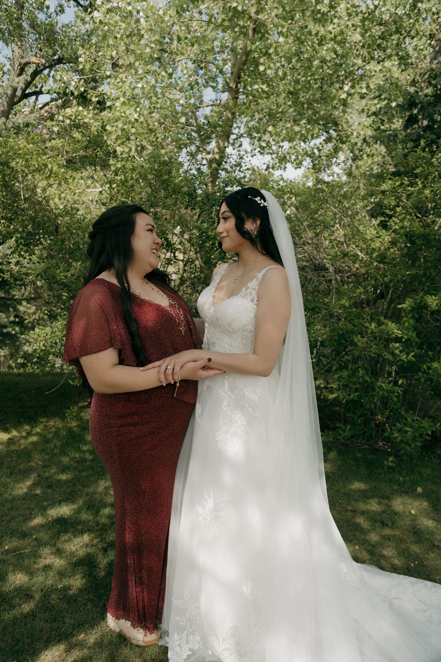 Bride in white lace gown and veil holds hands with woman in red dress, smiling at each other outdoors.