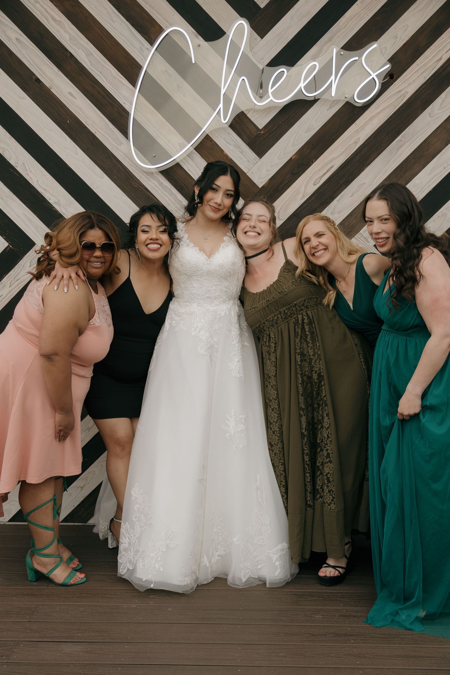 Bride in lace gown poses with five female guests at Cheers wedding photo backdrop.