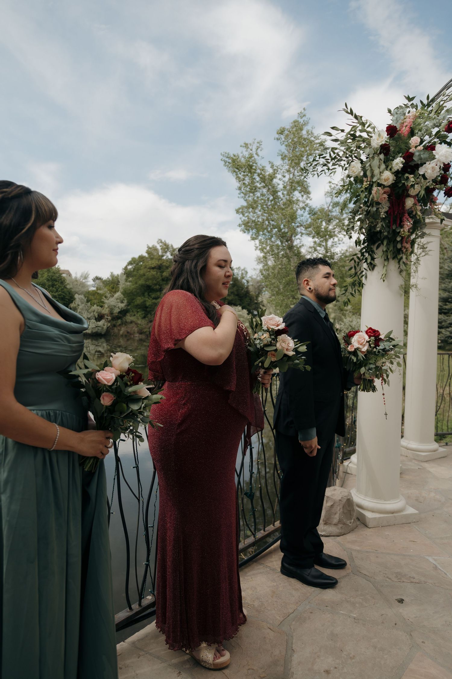 Bridesmaids in burgundy and sage dresses holding floral bouquets at outdoor wedding altar.