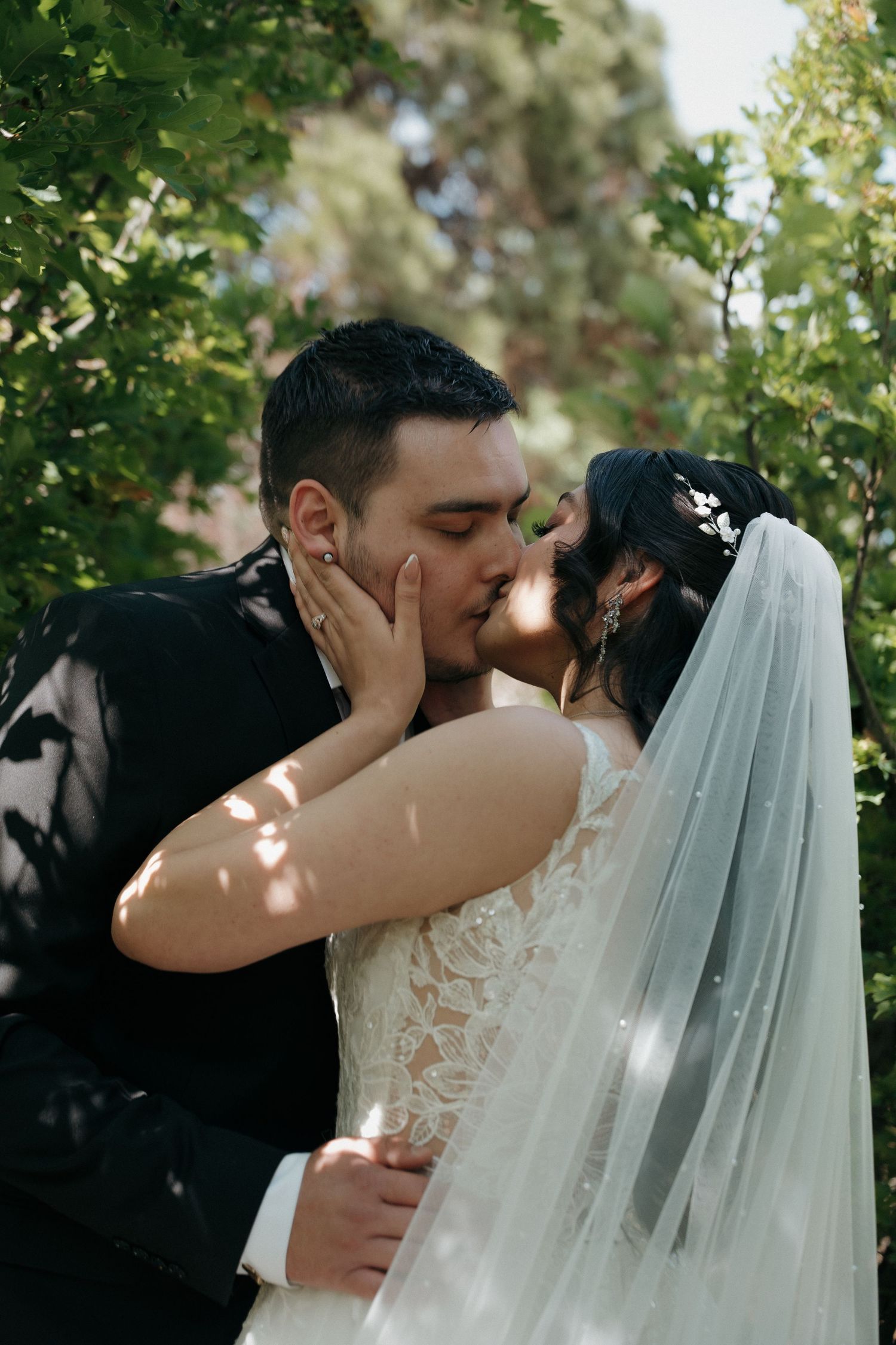 Bride and groom sharing a tender kiss surrounded by green foliage outdoors.