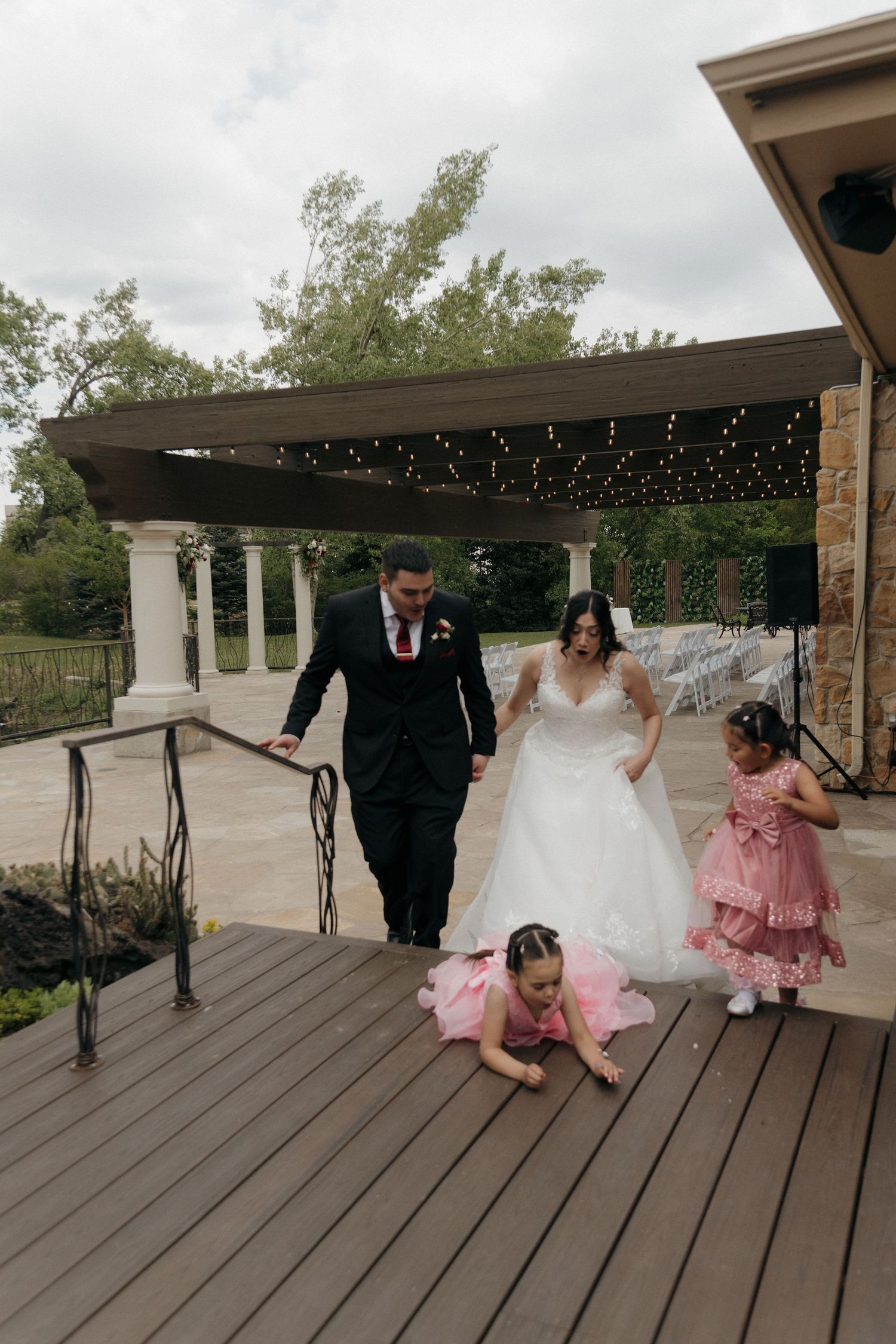 Bride and groom walk with flower girls on wooden deck after outdoor wedding ceremony on cloudy day.