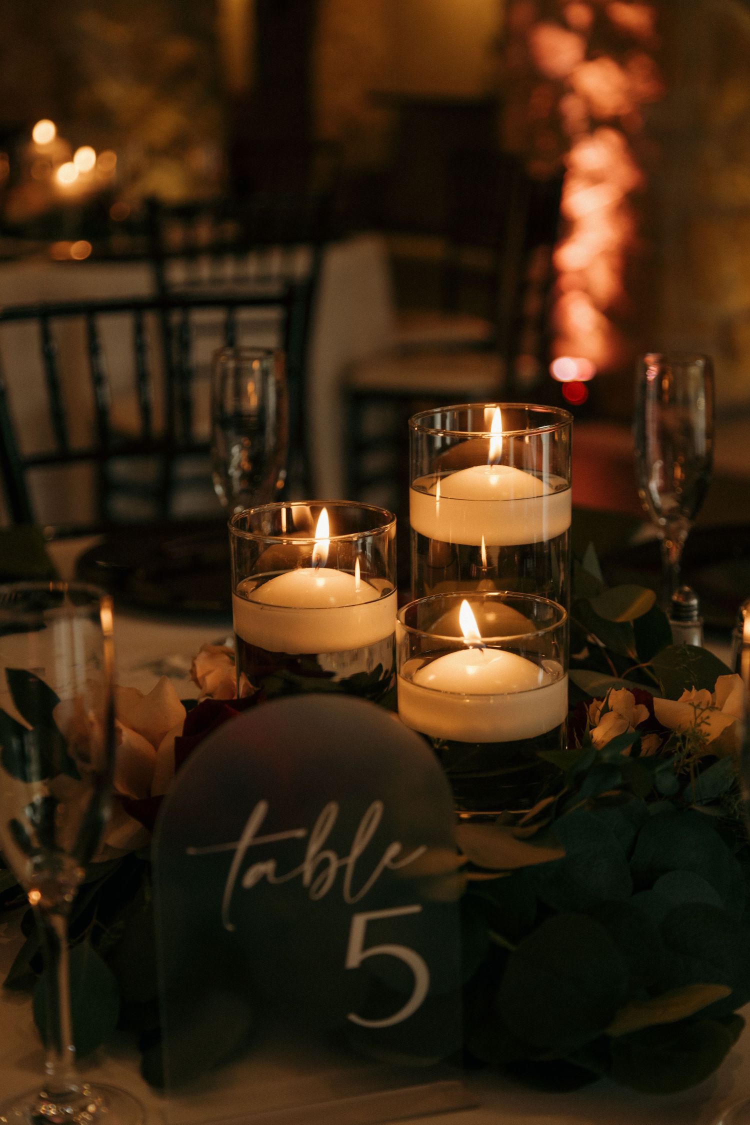 Elegant wedding table centerpiece with floating candles in glass cylinders and dark green table number 5 sign.