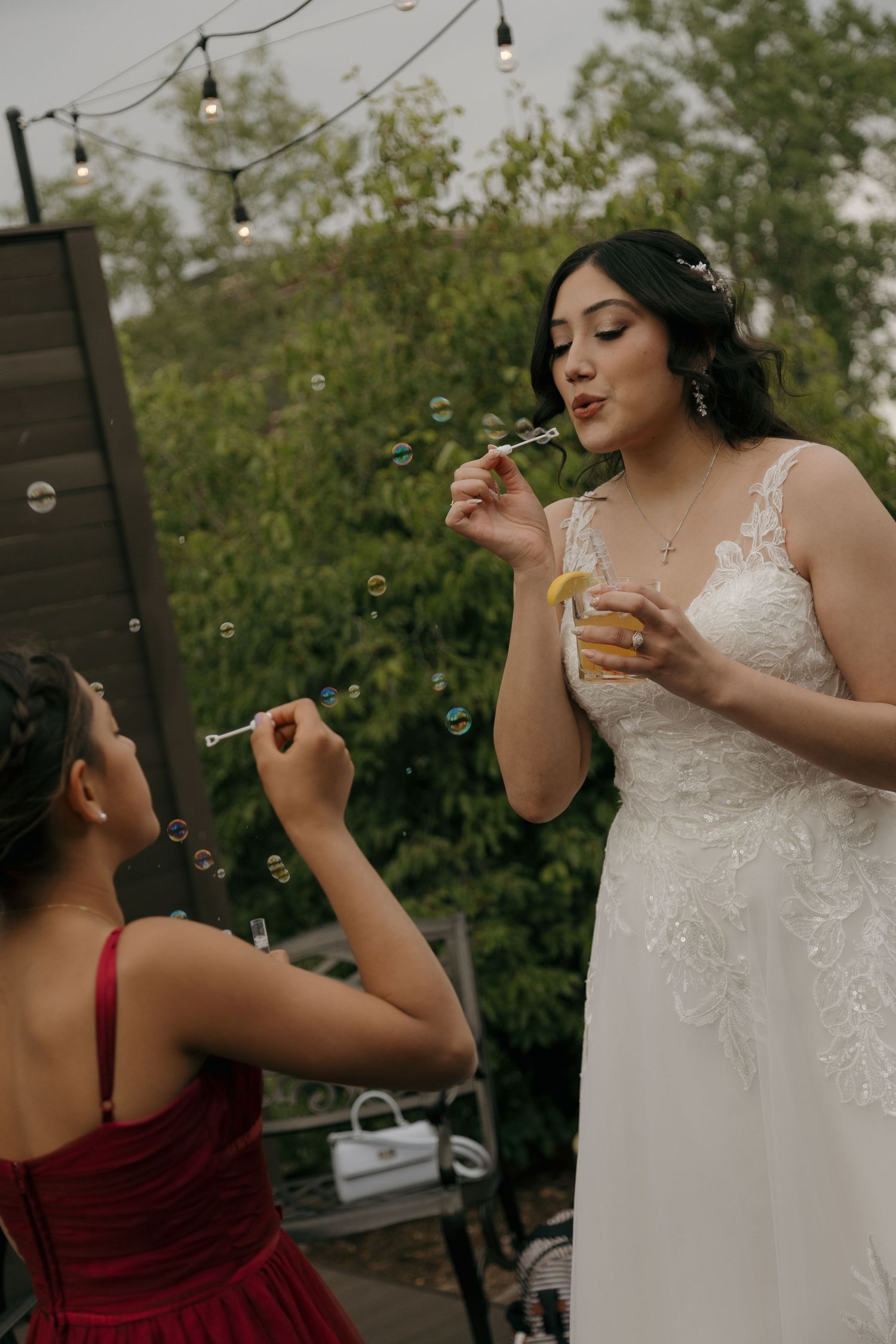 Bride in white lace gown blowing bubbles with bridesmaid in red dress at outdoor wedding reception.