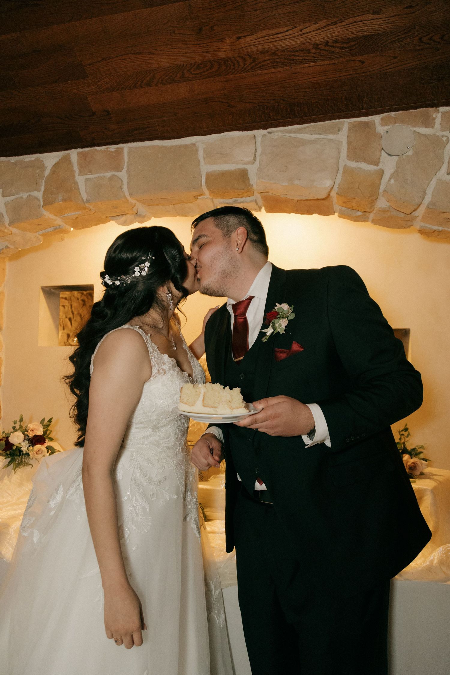 Bride and groom share a kiss while holding wedding cake slice in warmly lit stone arch venue.