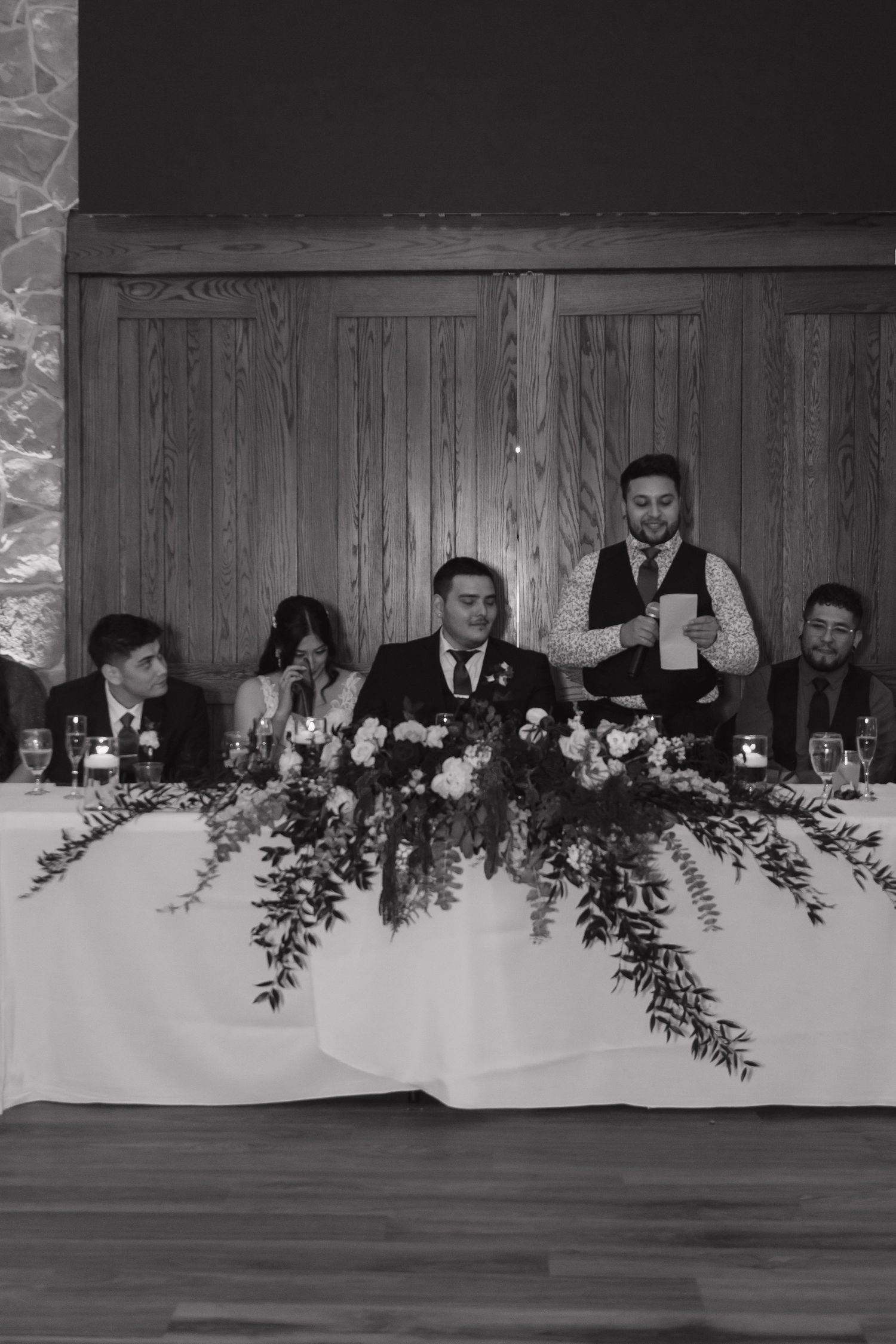 Black and white photo of wedding party at head table during best man speech at reception.