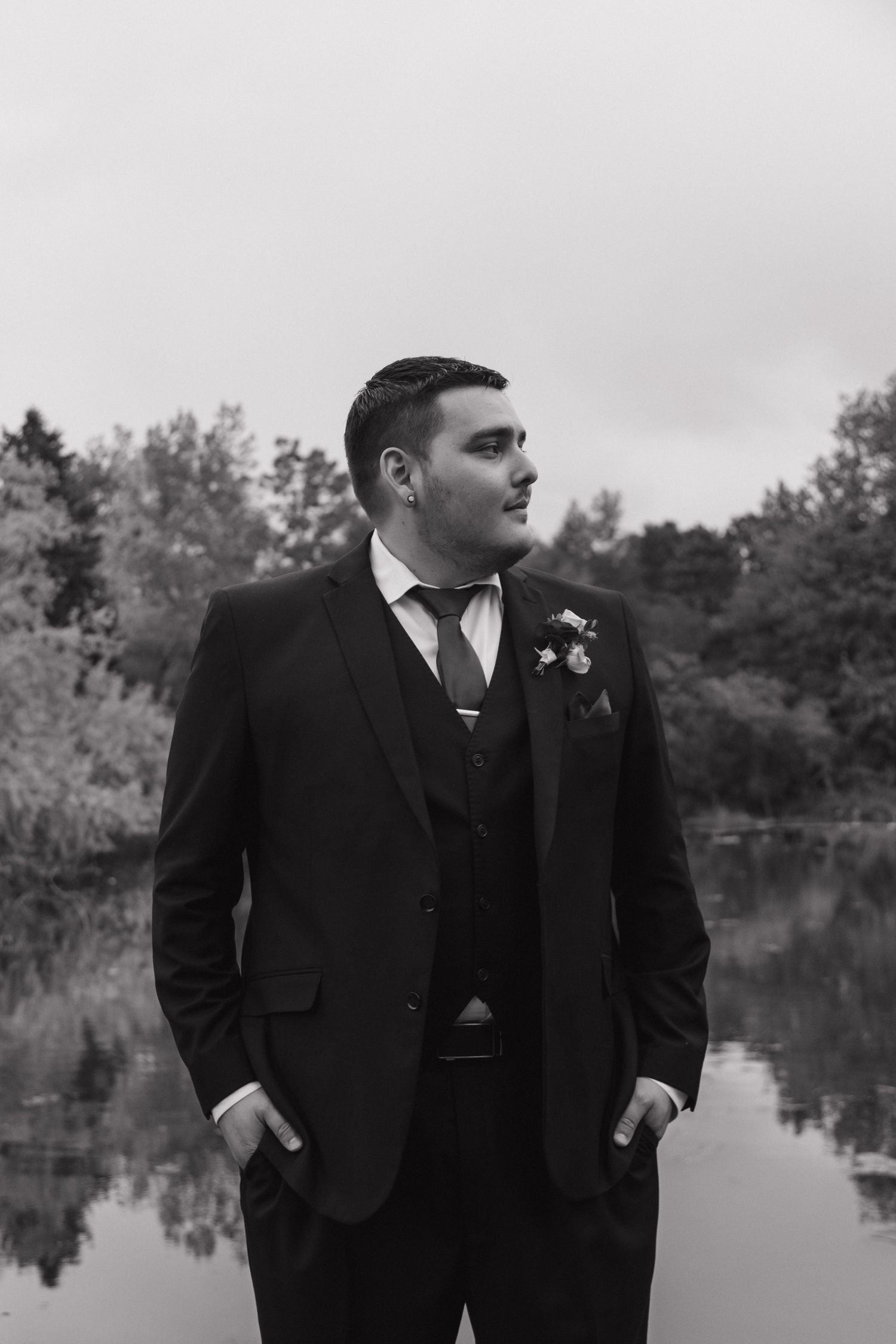 Groom in black three-piece suit with boutonniere poses by reflective lake in black and white photo.