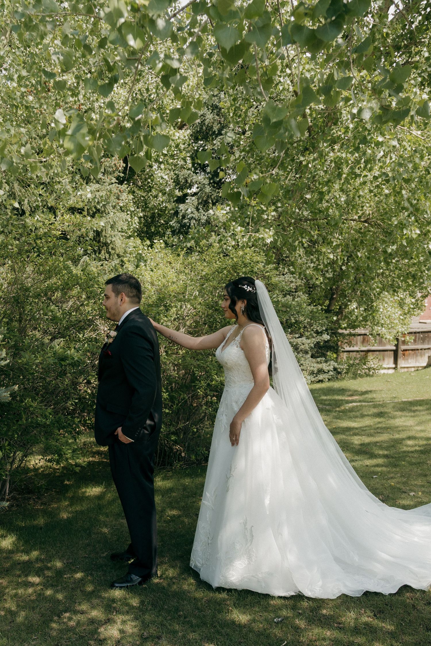 Bride in a white gown with veil taps groom on shoulder during outdoor first look under green trees.