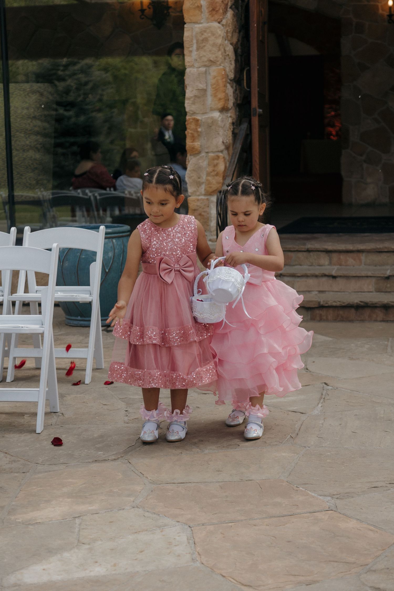 Two little flower girls in pink dresses scatter rose petals along the aisle at an outdoor wedding ceremony.