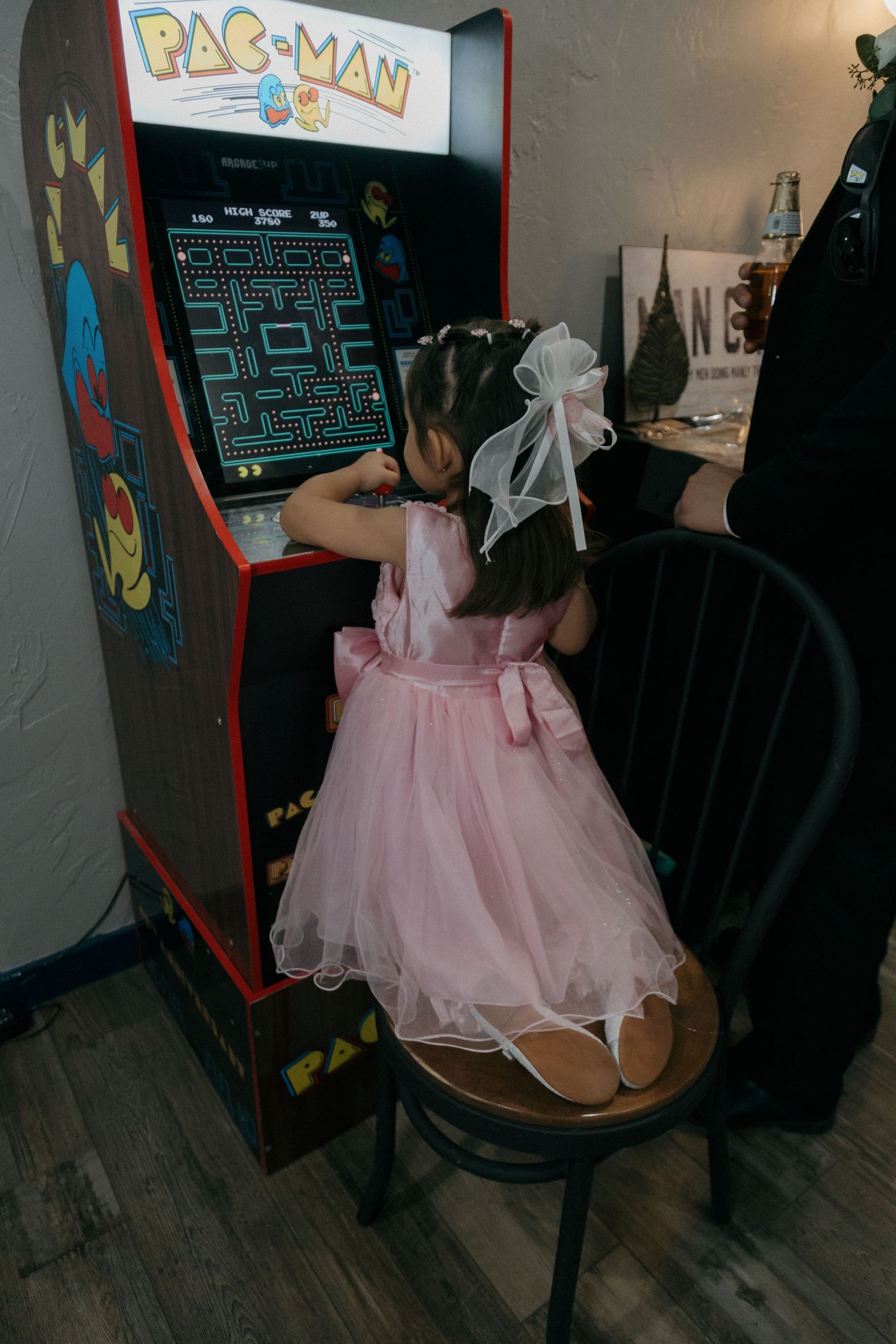 Young girl in pink dress plays Pac-Man arcade game standing on chair at wedding reception.