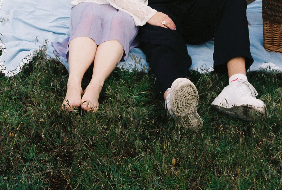 Two people sitting on grass, one barefoot in a purple skirt, the other wearing white sneakers on a sunny day.