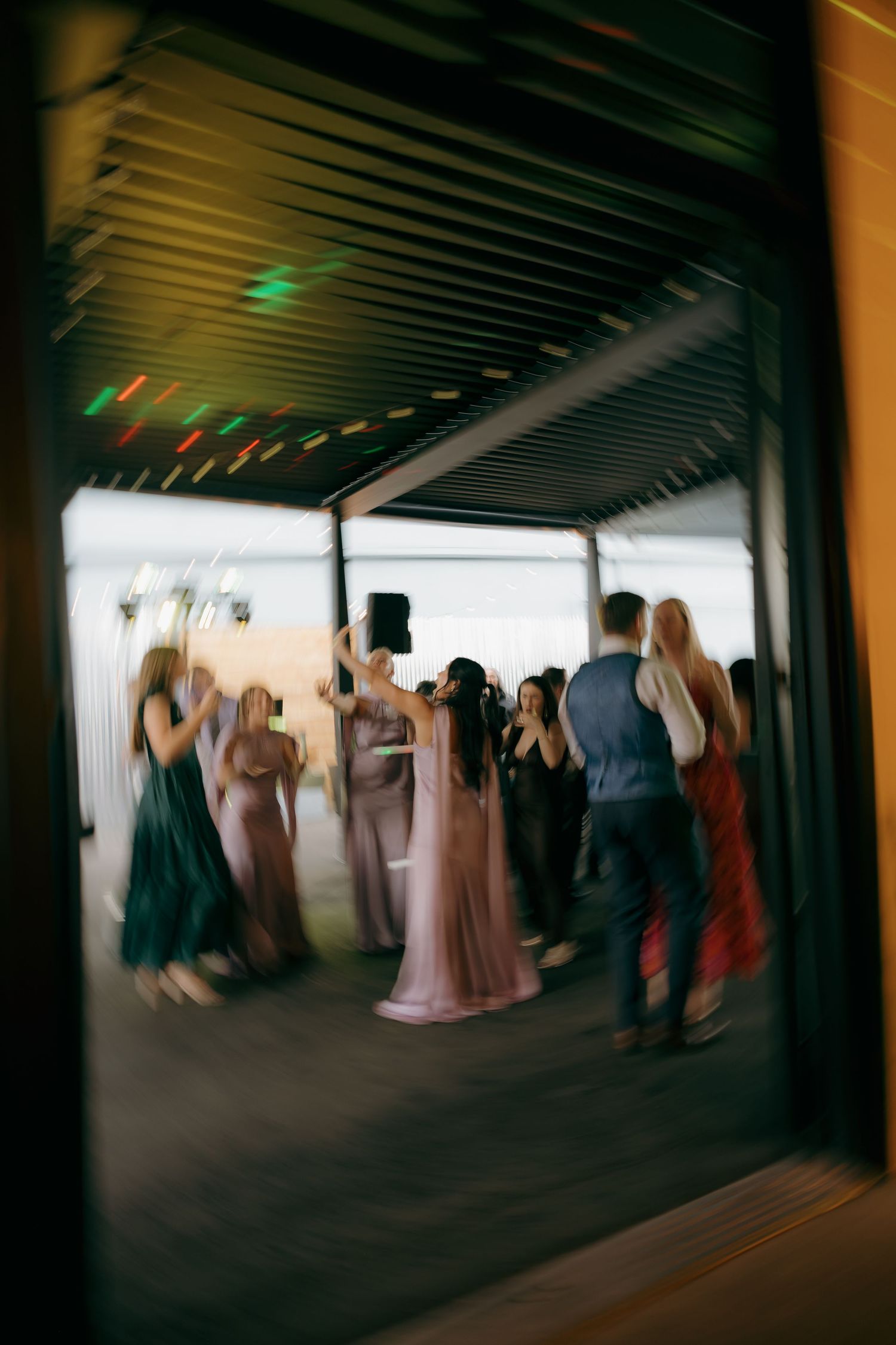 Wedding guests dance energetically at a reception, captured with artistic motion blur through a glass door.