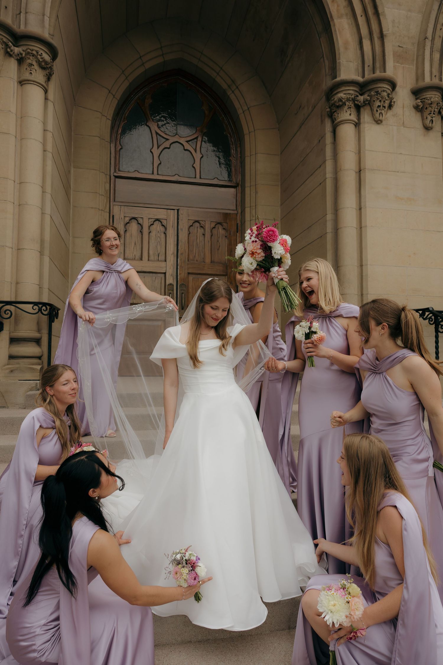 Bridesmaids in purple gowns surround bride in white dress on stone church steps, holding colorful bouquets.