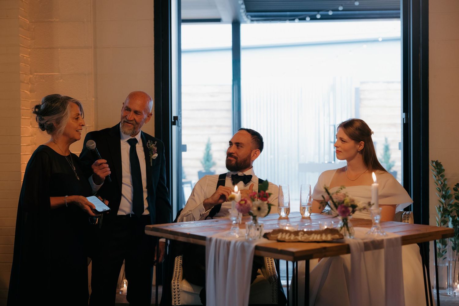 Parents giving toast speech to newlyweds seated at sweetheart table during reception.