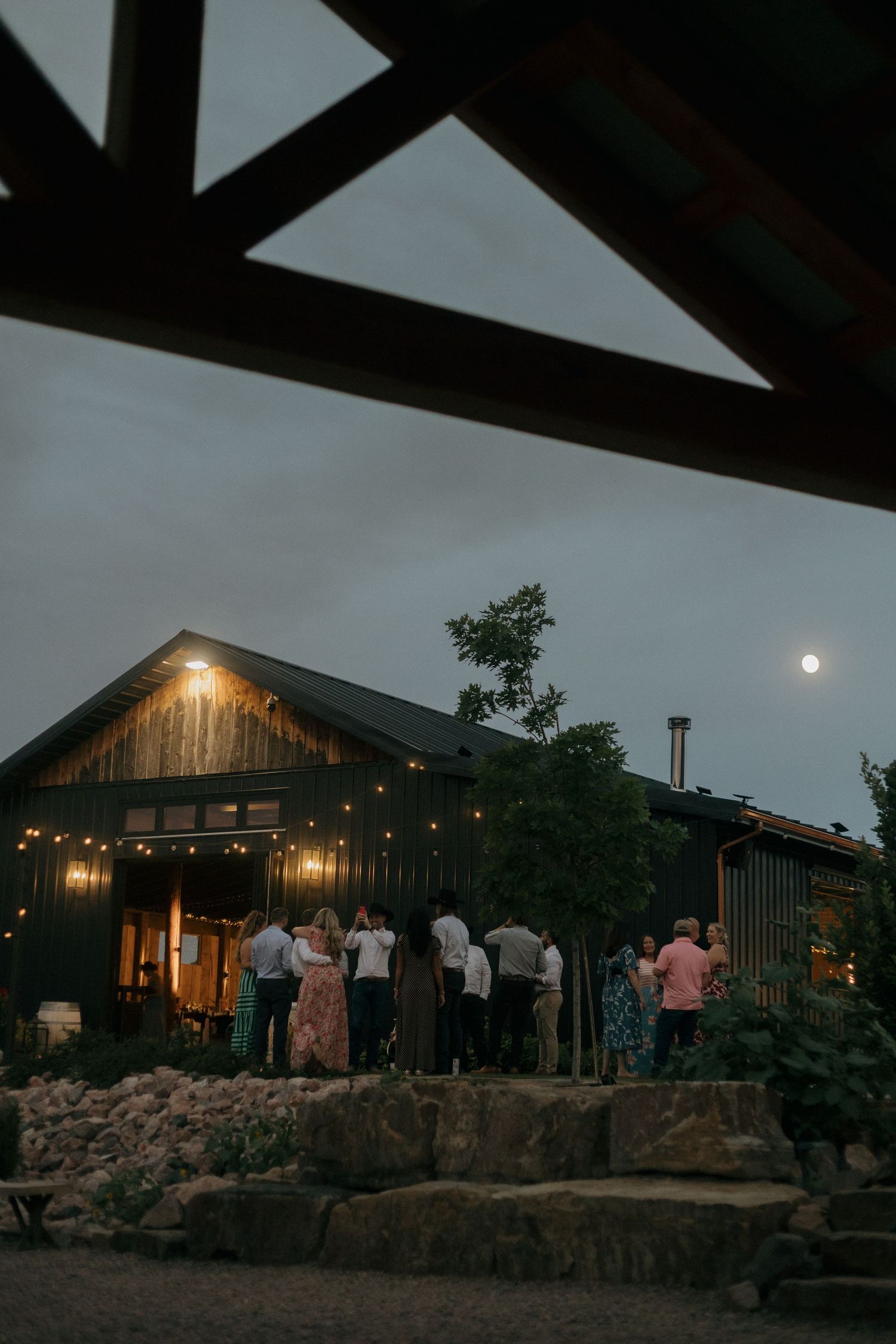 Guests gather outside a dark barn venue decorated with string lights under a moonlit evening sky.