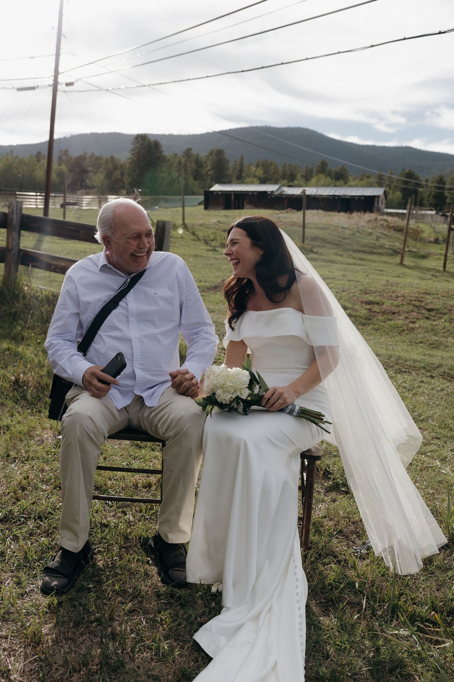 Bride in white gown and veil laughs with elderly man in white shirt, seated outdoors at rustic farm wedding venue.