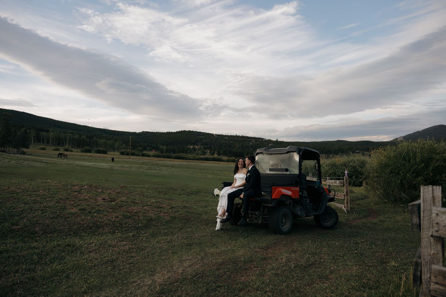 Bride and groom sitting together on an orange utility vehicle in a scenic mountain meadow.