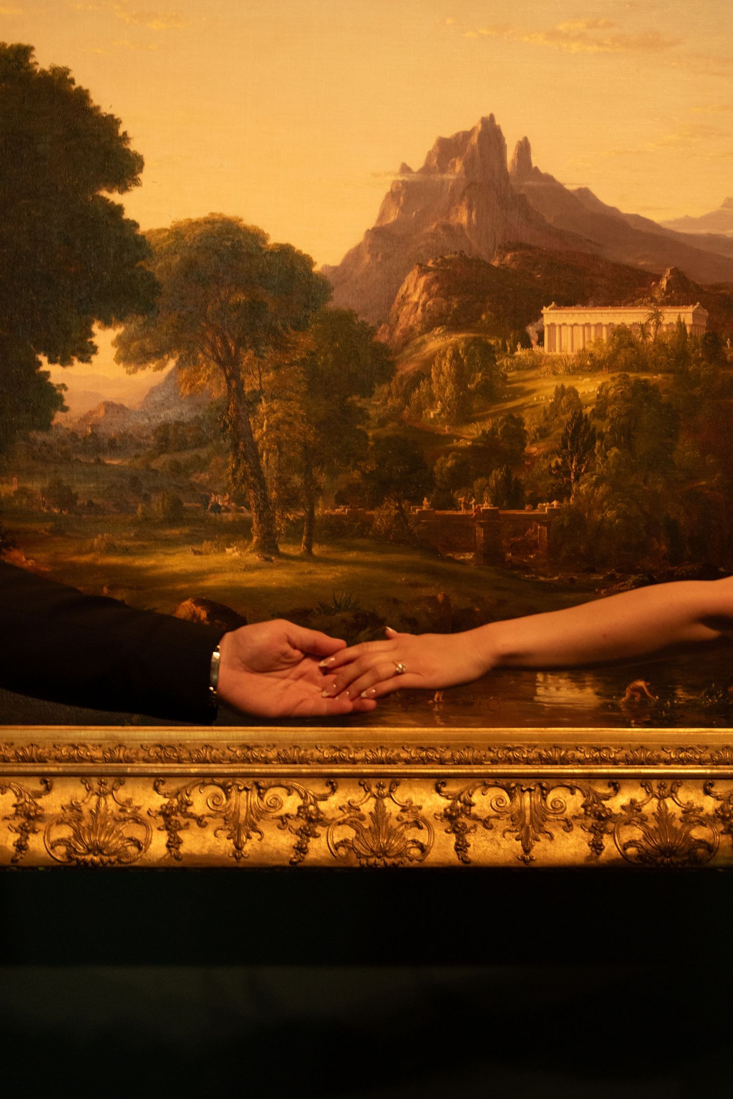 Hands reach across a romantic landscape painting with mountains and trees in an ornate gold frame.