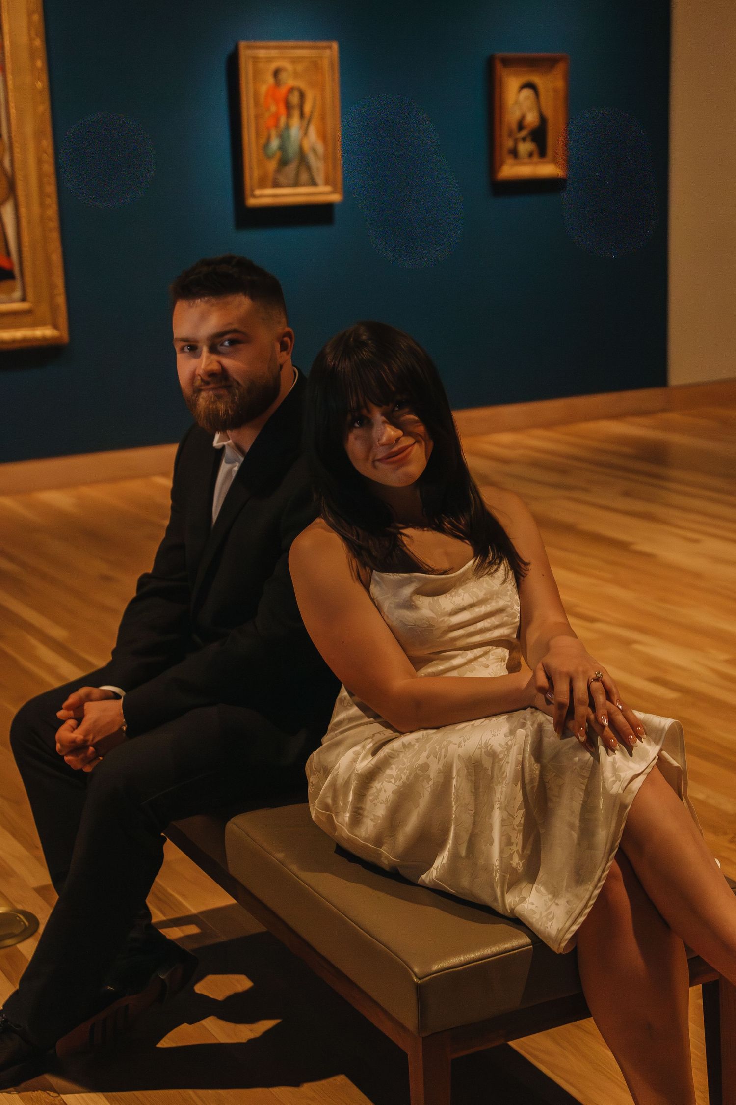 A couple sits together on a bench in an art museum with blue walls.