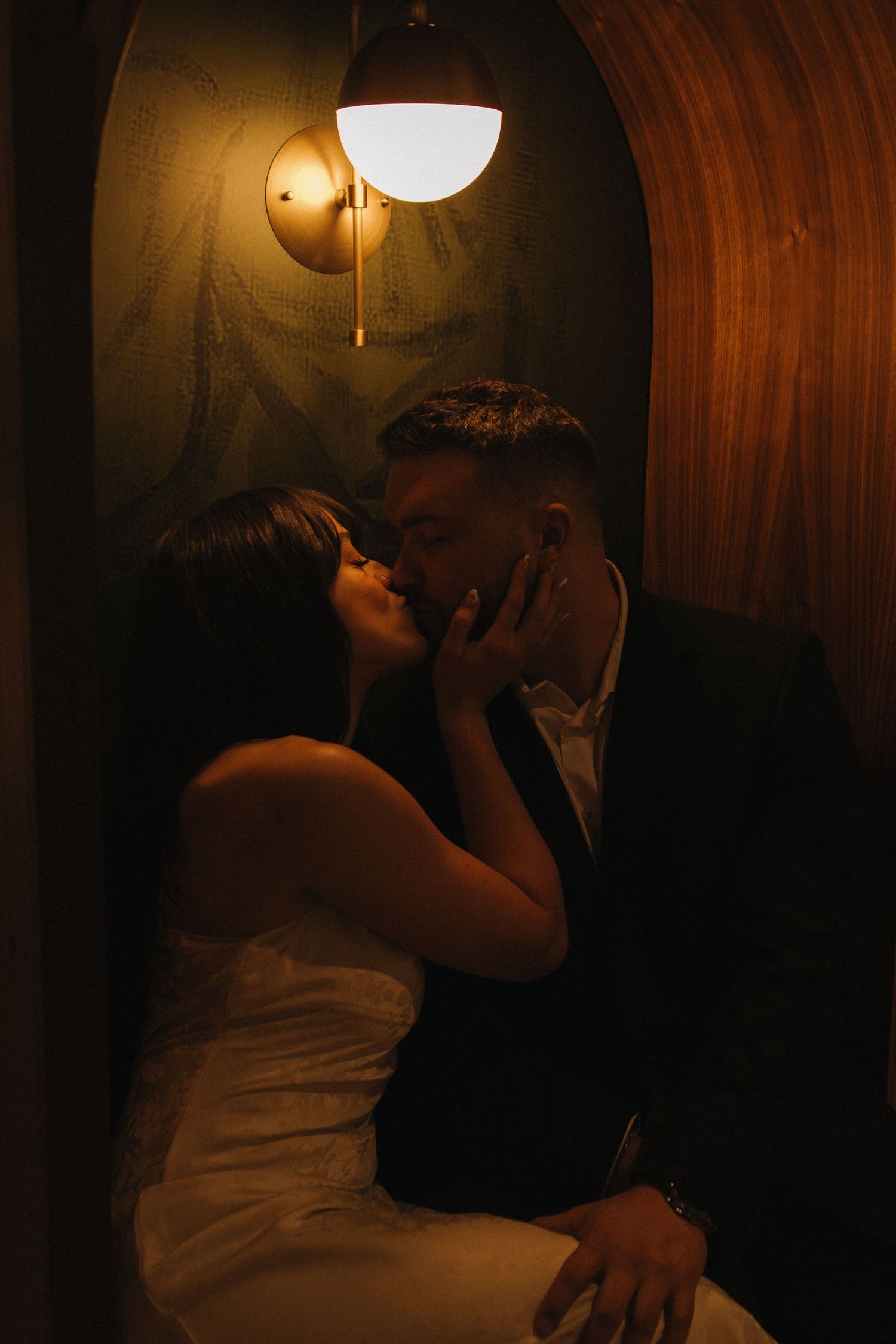 Romantic couple embracing in dimly lit alcove with art deco wall sconce.