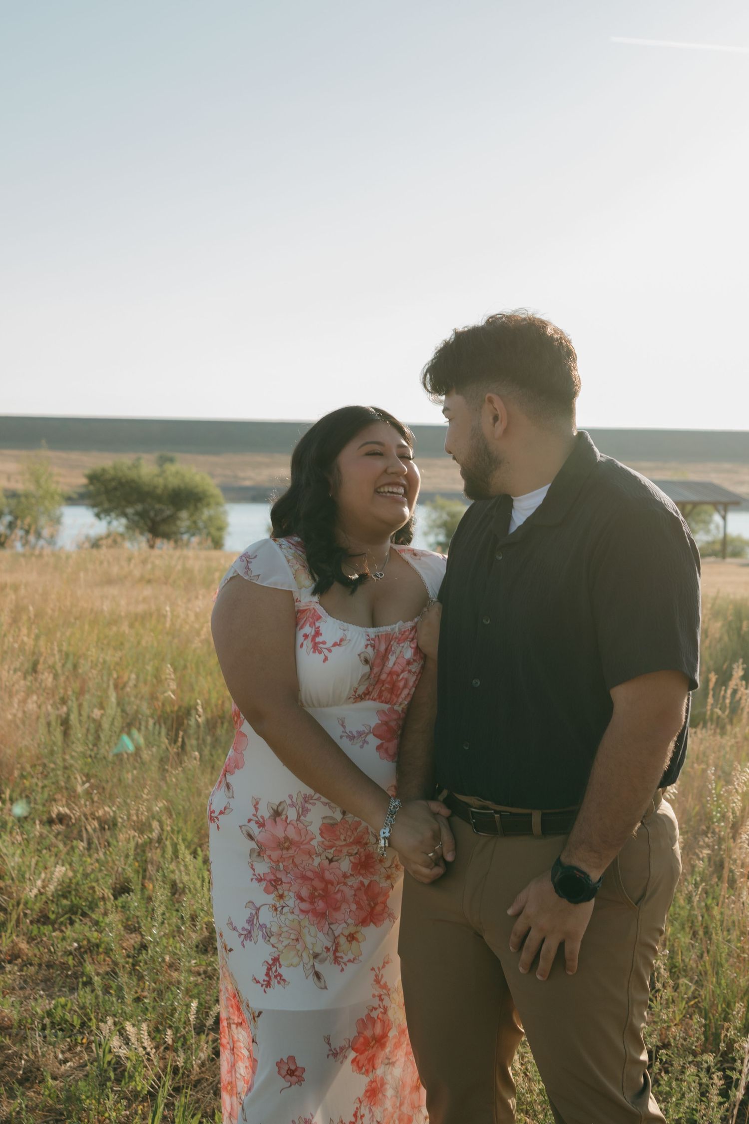 A couple shares a joyful moment together in a field with a lake and hills visible in the background.