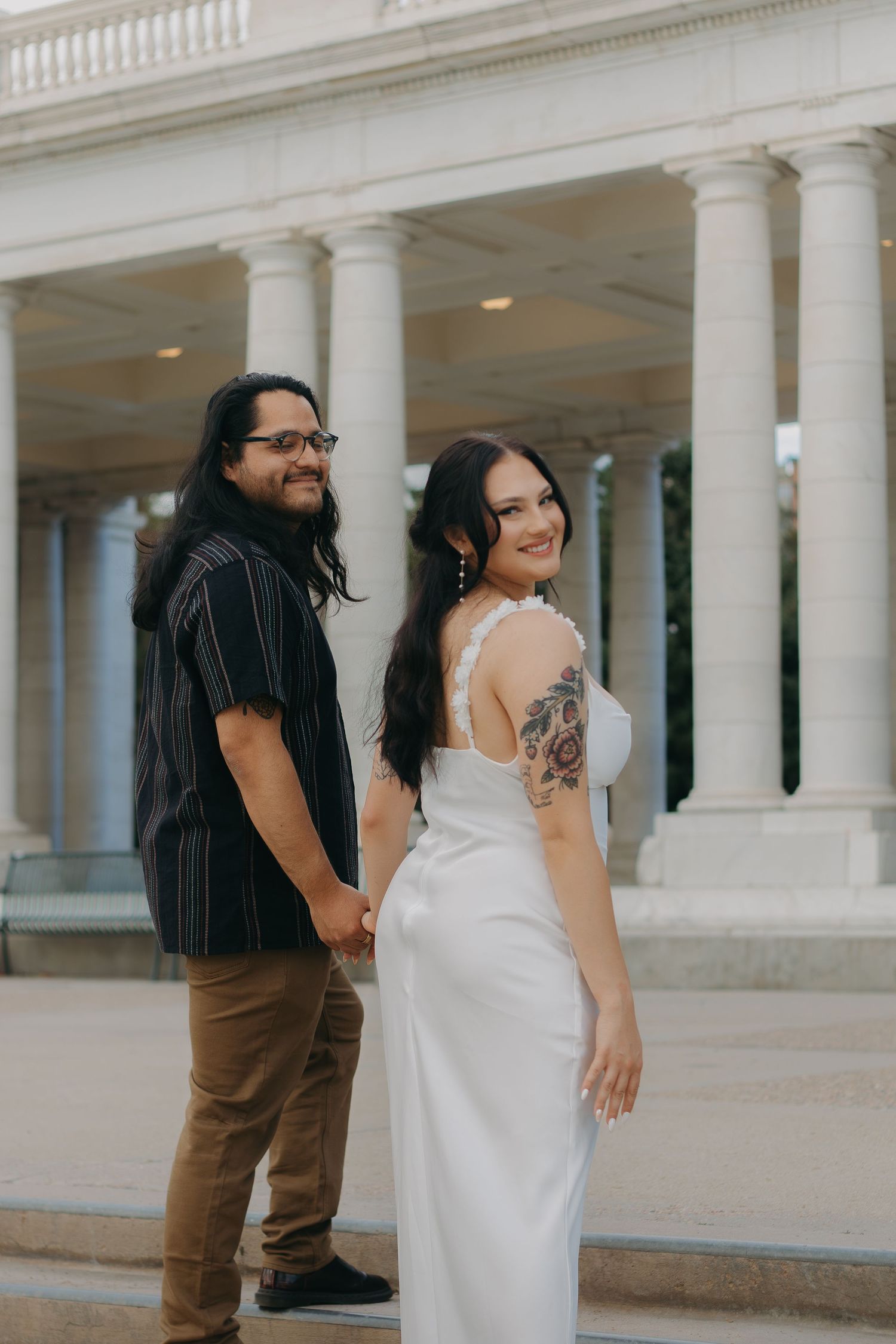 Couple poses together in front of elegant white columned building, with bride wearing tattoo-revealing wedding gown.