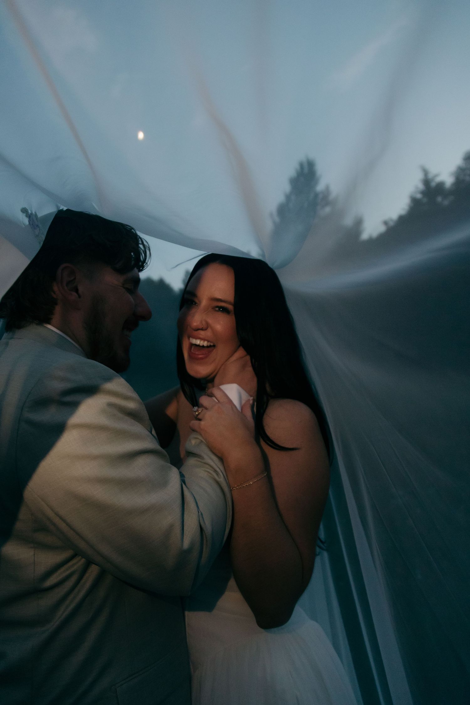 Joyful bride laughs with groom under flowing wedding veil in soft blue twilight forest setting.