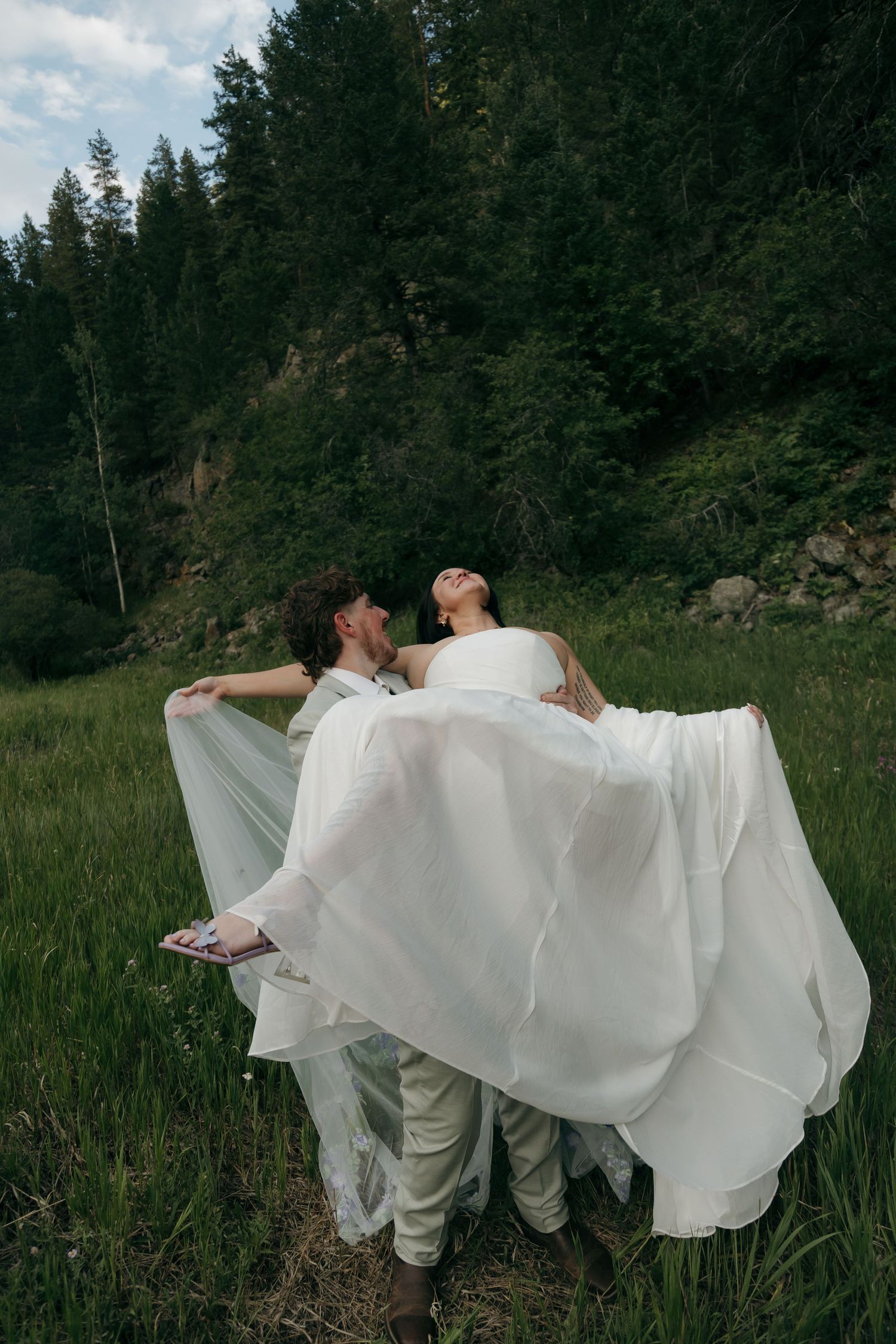 Groom lifting bride in white wedding dress outdoors in a green forest meadow setting.