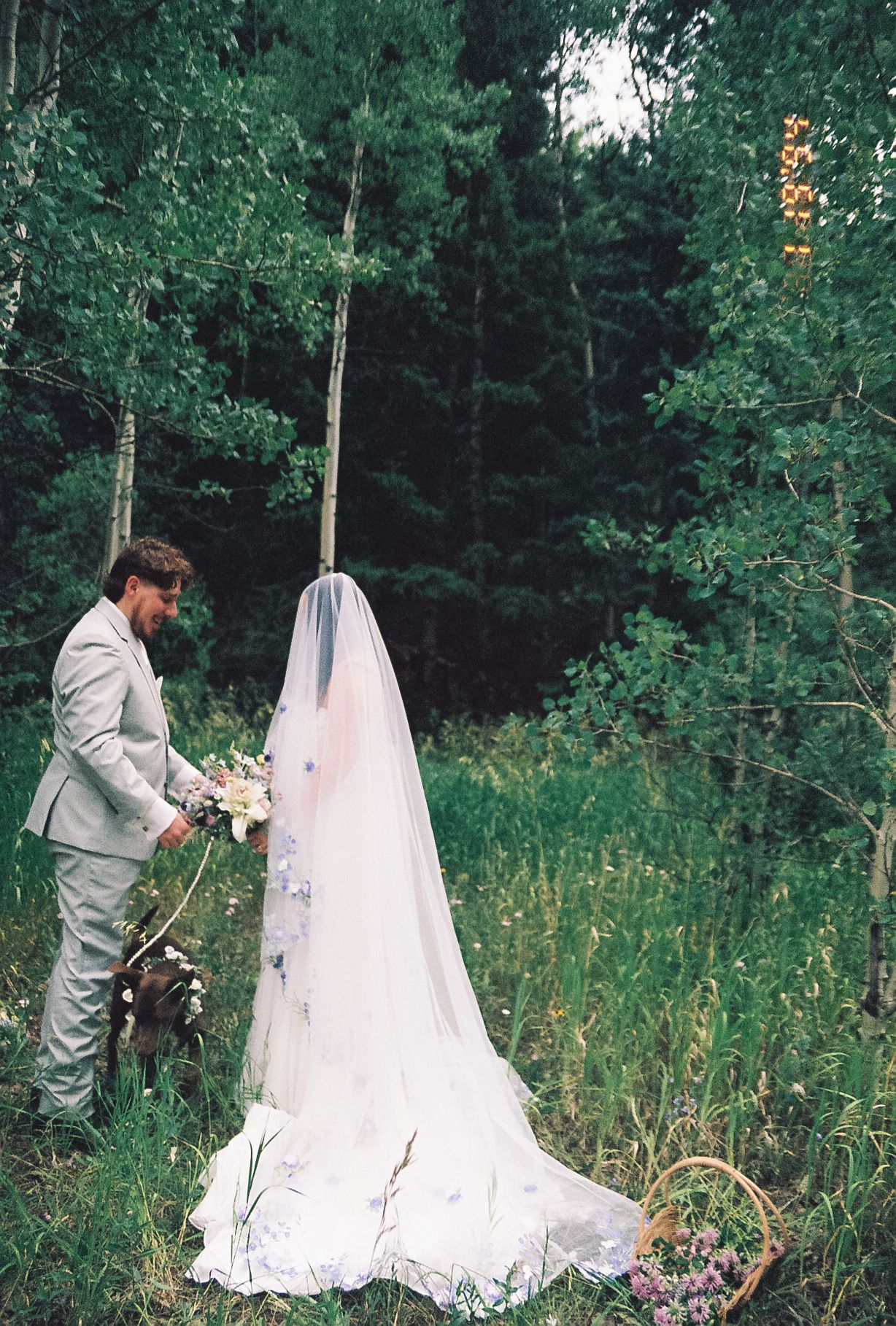 Groom in gray suit holds bride's bouquet as she stands in a long veil in a green forest during their outdoor elopement.