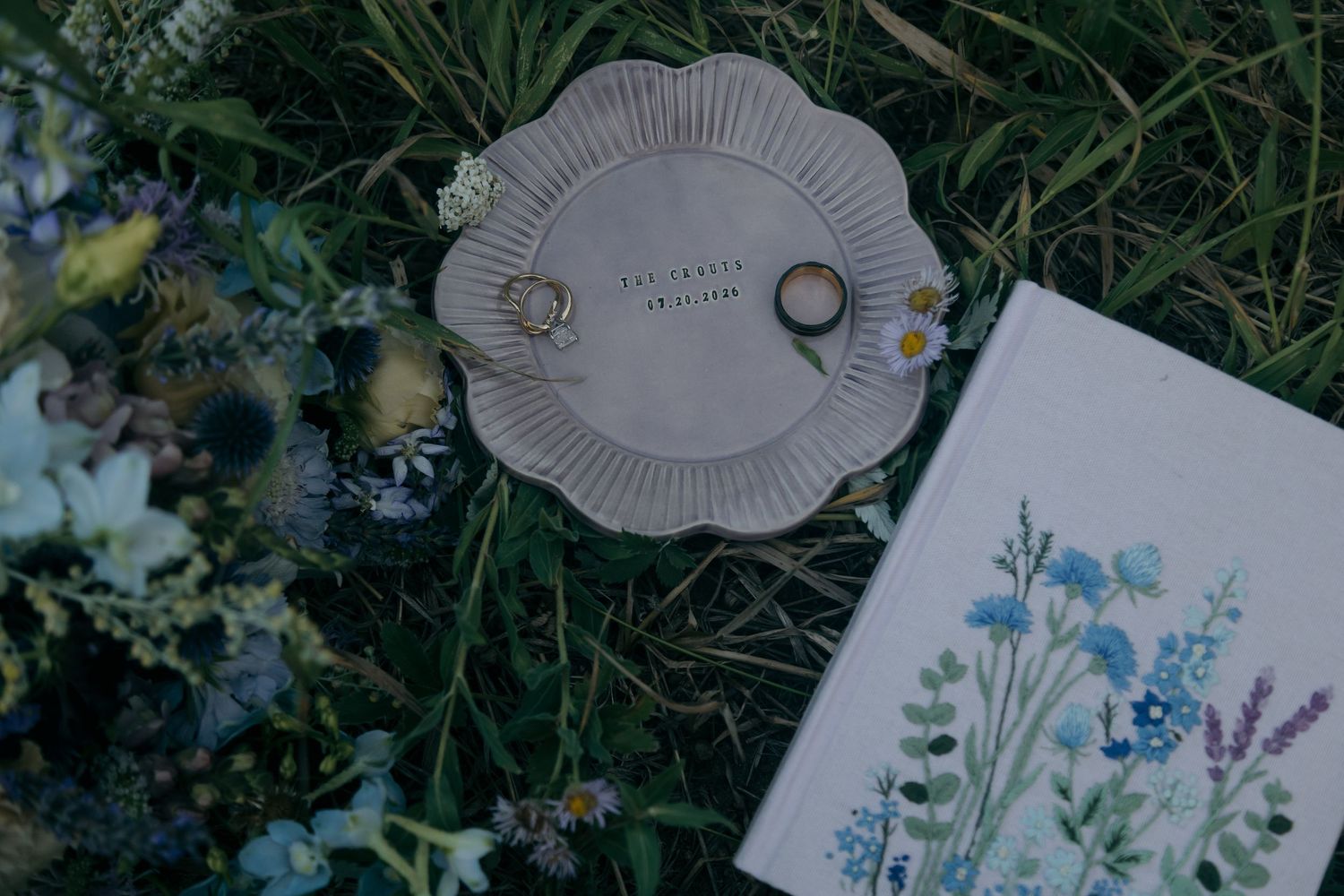 Flat lay of wedding rings on ceramic dish beside floral vow book and wildflowers in grass.