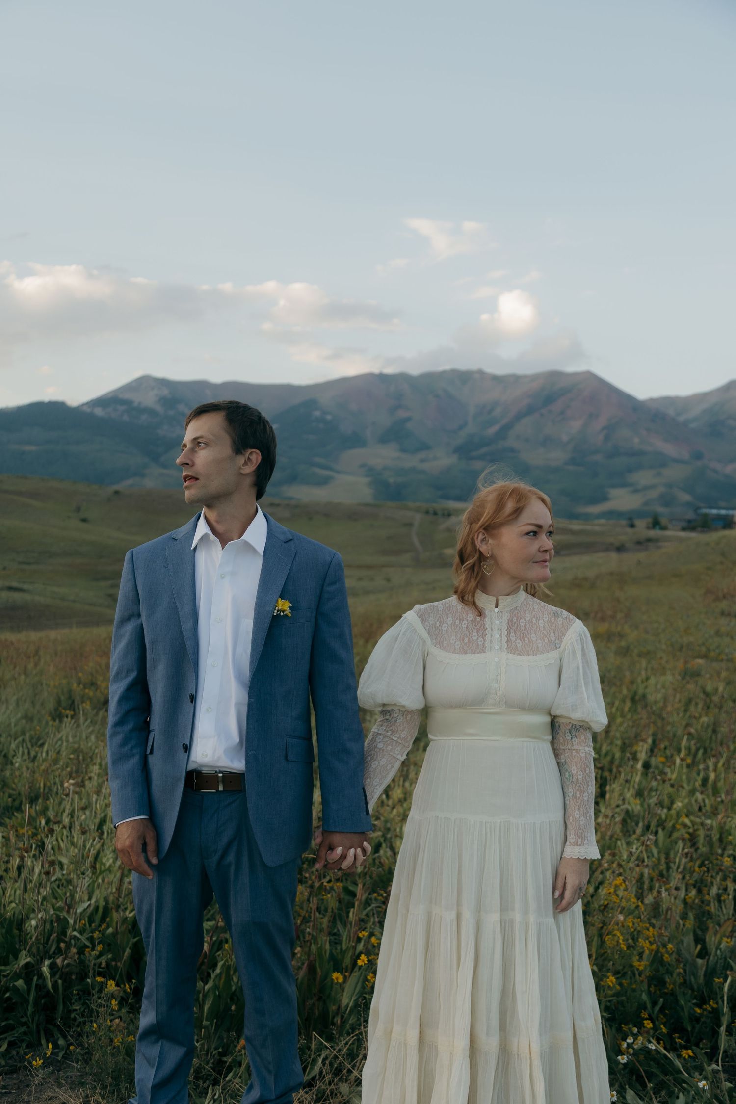 Bride and groom holding hands in mountain meadow, wearing blue suit and vintage lace wedding dress.