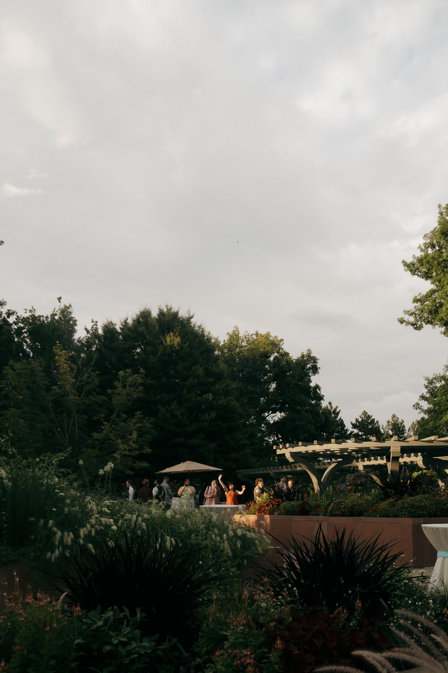 Wedding guests mingling outdoors in a lush garden venue with a wooden pergola under a cloudy evening sky.
