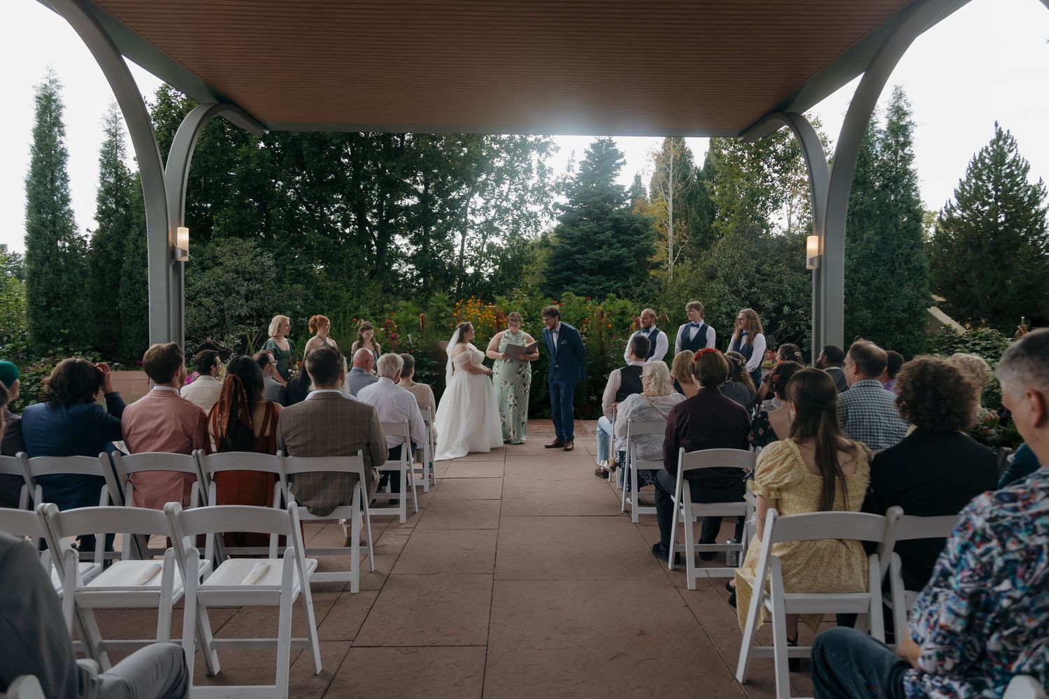 Outdoor wedding ceremony under a pergola with guests seated in white chairs surrounded by lush greenery.