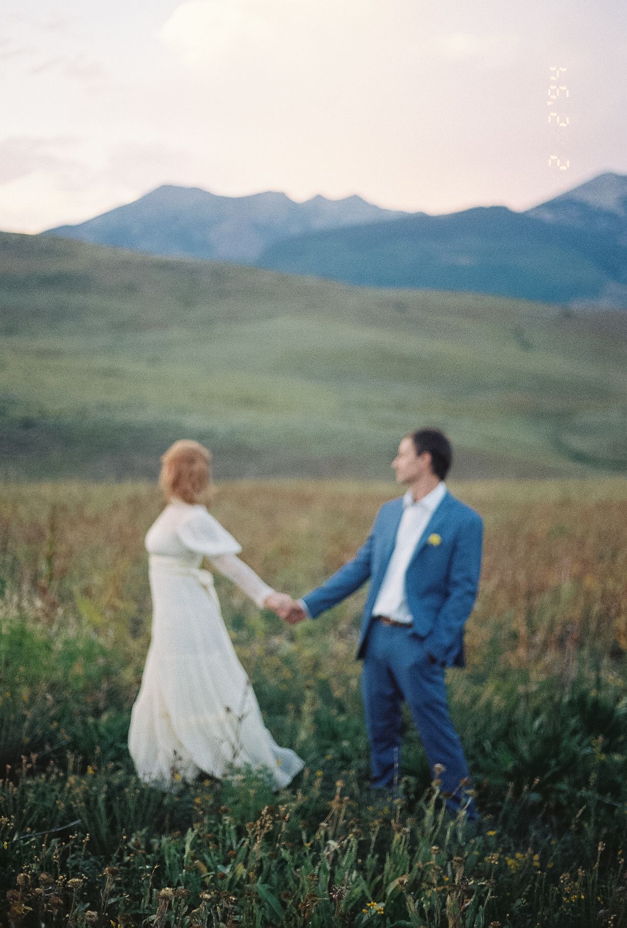 Bride and groom hold hands in a wildflower meadow with misty blue mountains in the background at dusk.