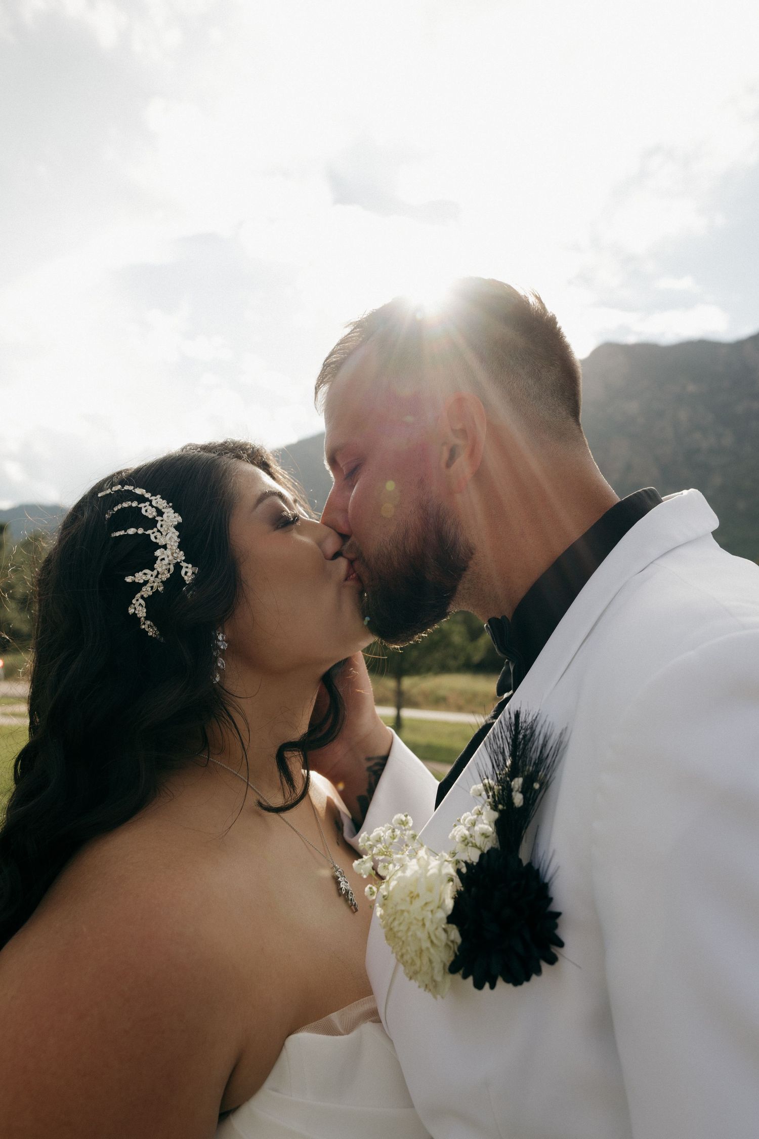 Bride and groom share passionate kiss with sun flare and mountain backdrop on their wedding day.