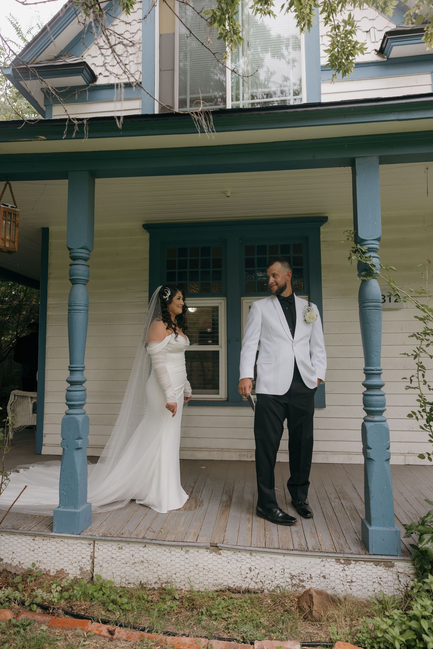 Bride in elegant white gown and groom in white jacket share tender moment on blue-trimmed porch.