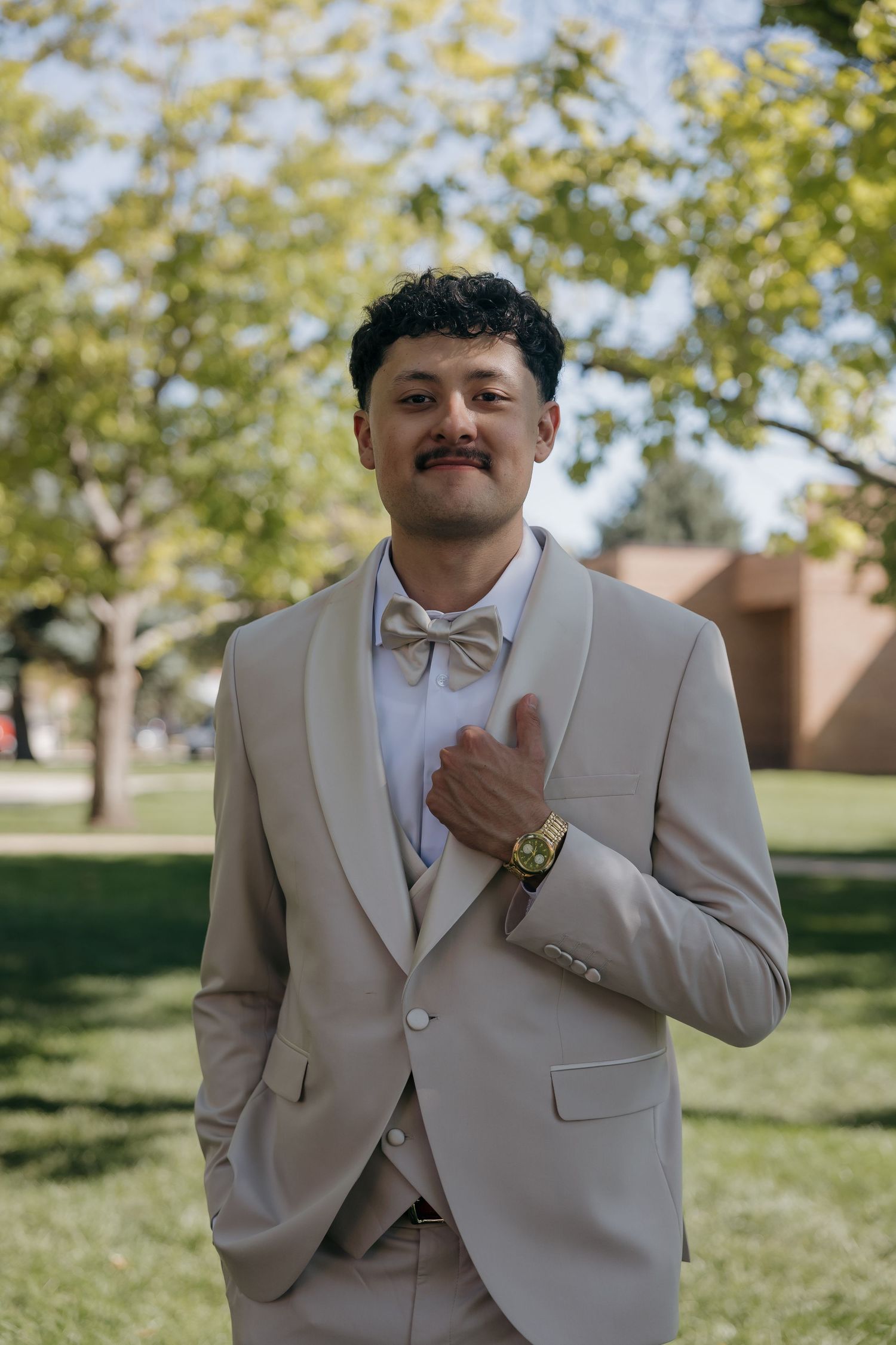 Young man in beige tuxedo with bow tie and gold watch smiling outdoors at a formal event.