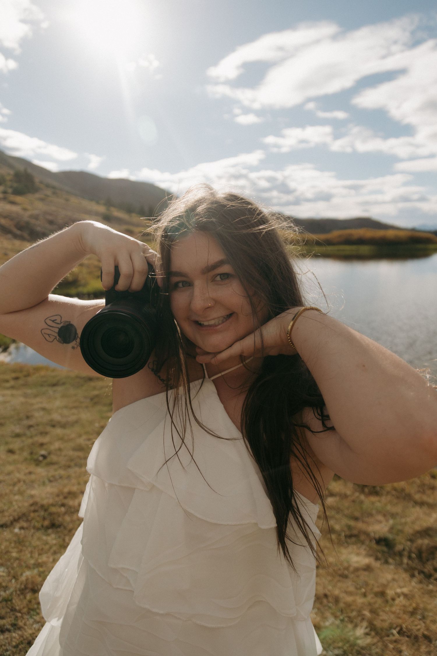 A smiling woman in a white dress holds a DSLR camera outdoors near a scenic mountain lake on a sunny day.